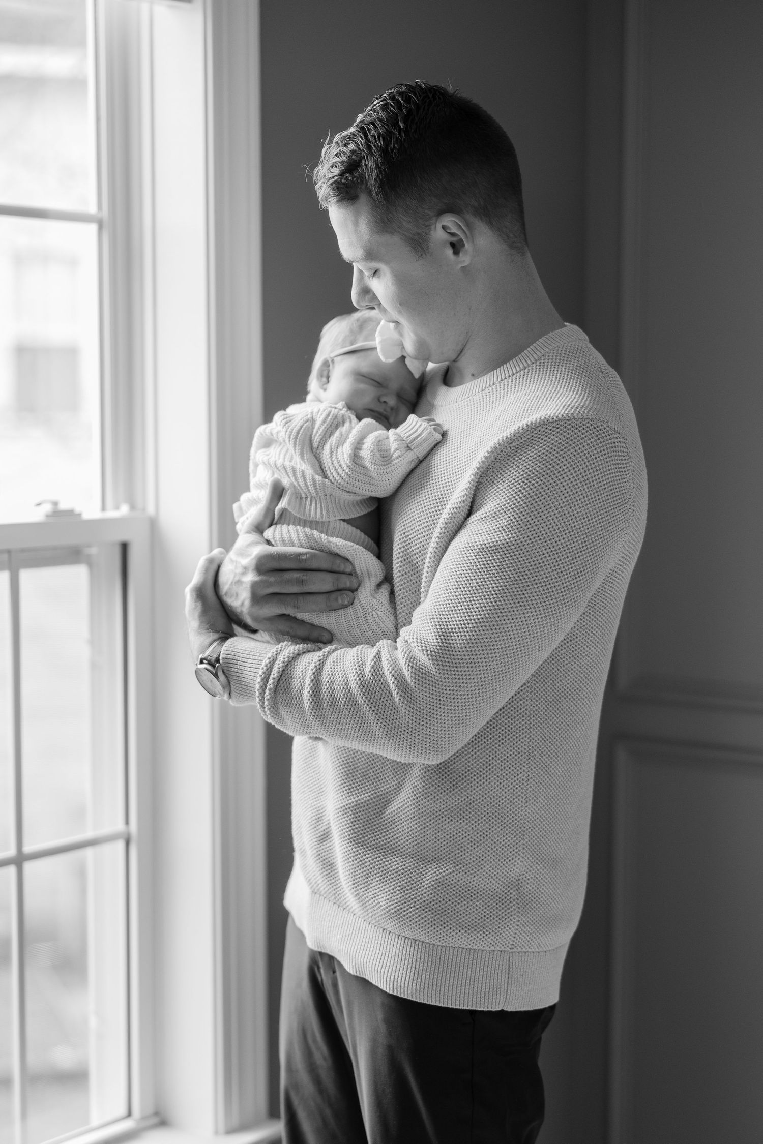 Black and white portrait of parent holding newborn baby near window.