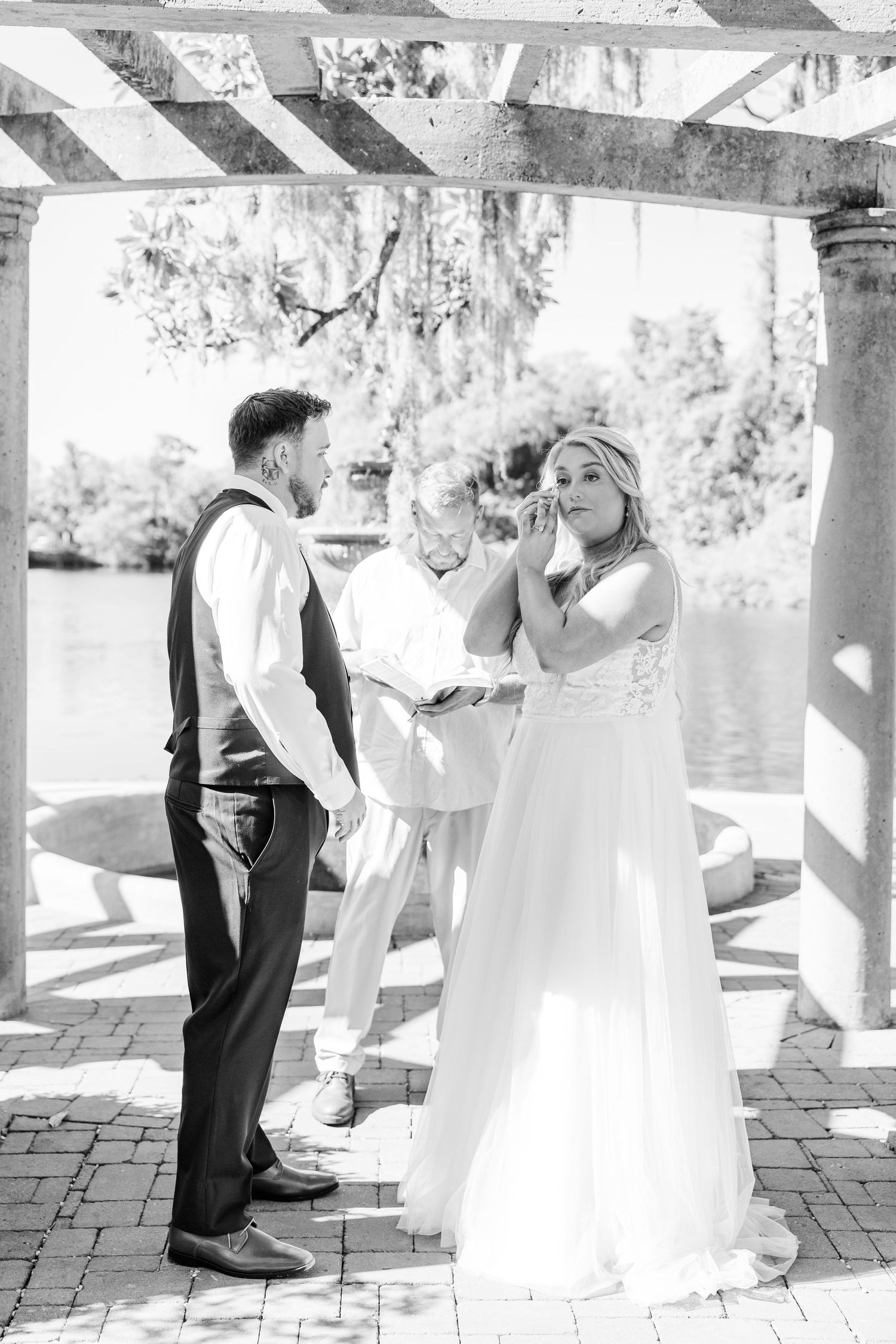 Black and white photo of emotional bride wiping tears during outdoor wedding ceremony with groom and officiant in Airlie Gardens in Wilmingt