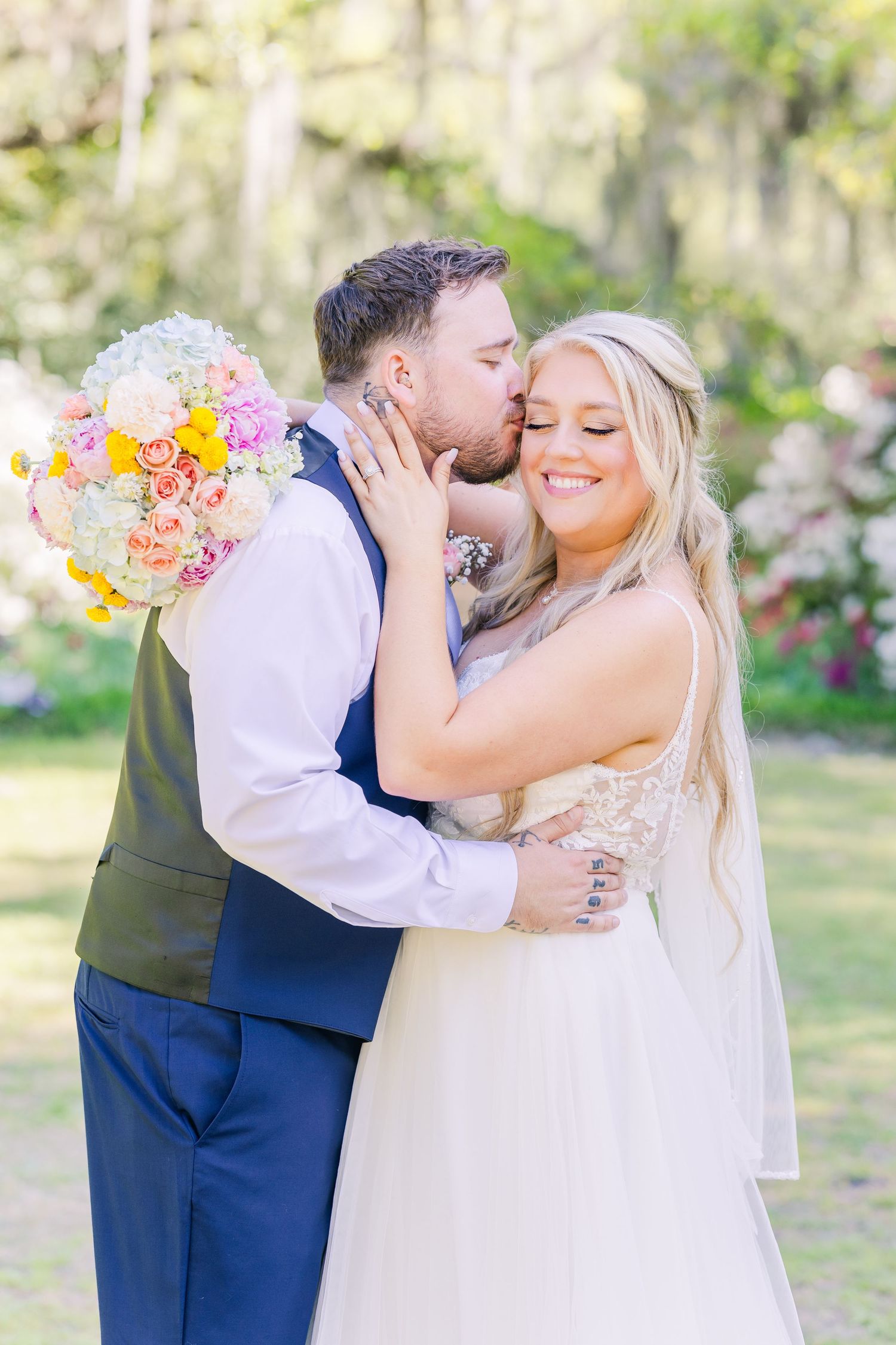 Groom kisses bride on cheek as she smiles holding colorful pink and yellow wedding bouquet outdoors in Airlie Gardens in Wilmington, NC