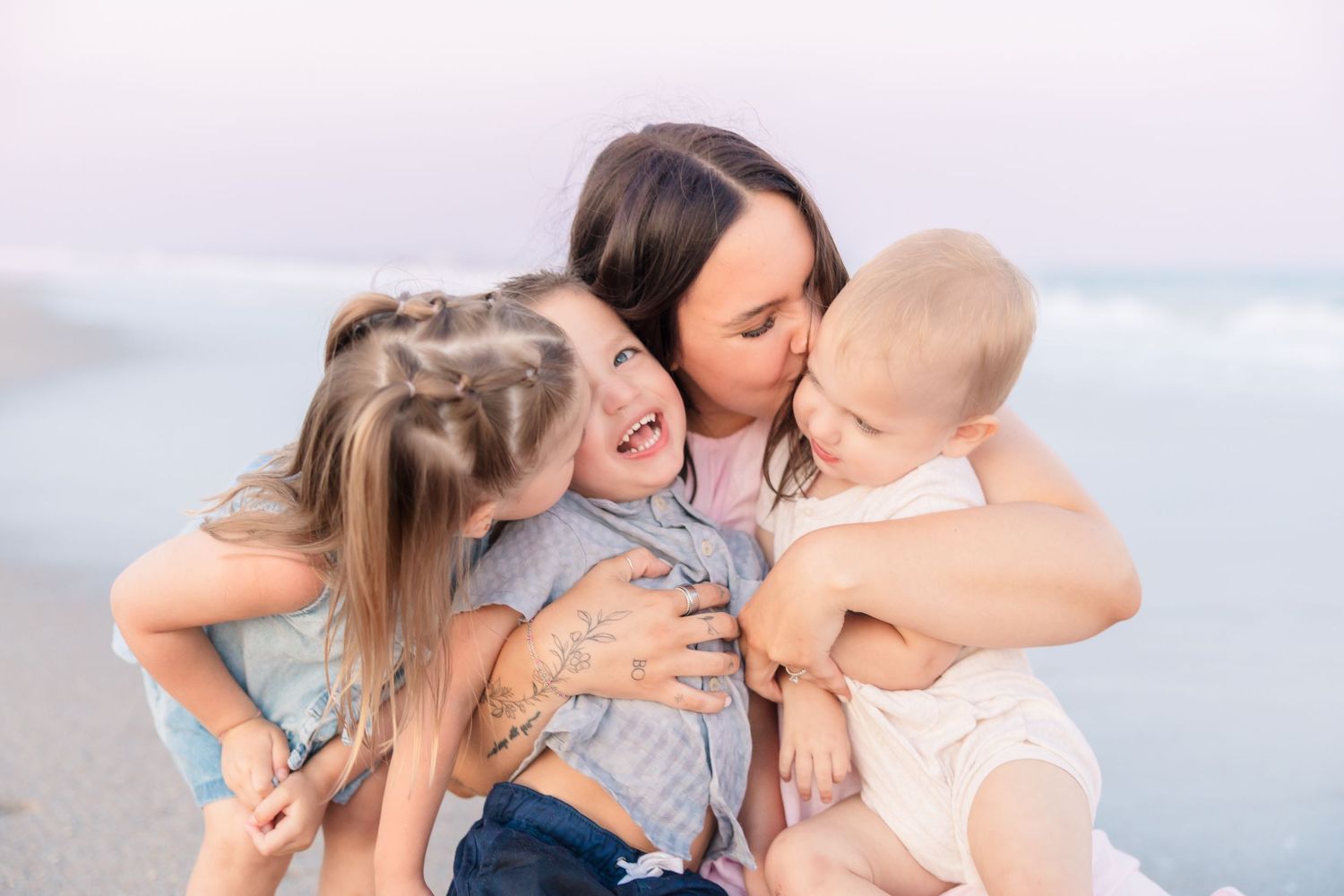 A mother sits on a beach hugging and kissing her three young children, sharing a joyful family moment by the ocean.