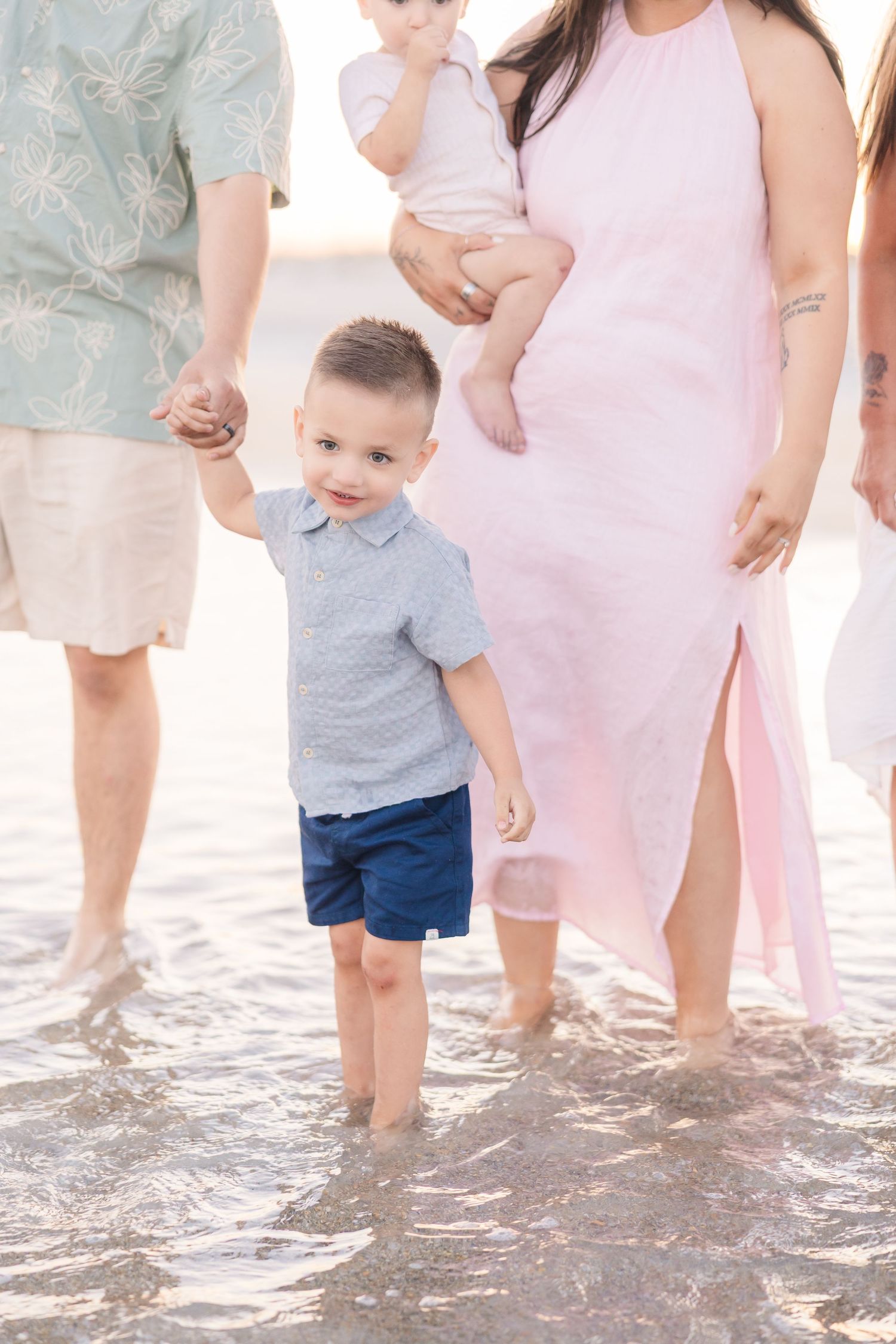 Young boy holds mothers hand while wading in shallow beach water with family nearby.