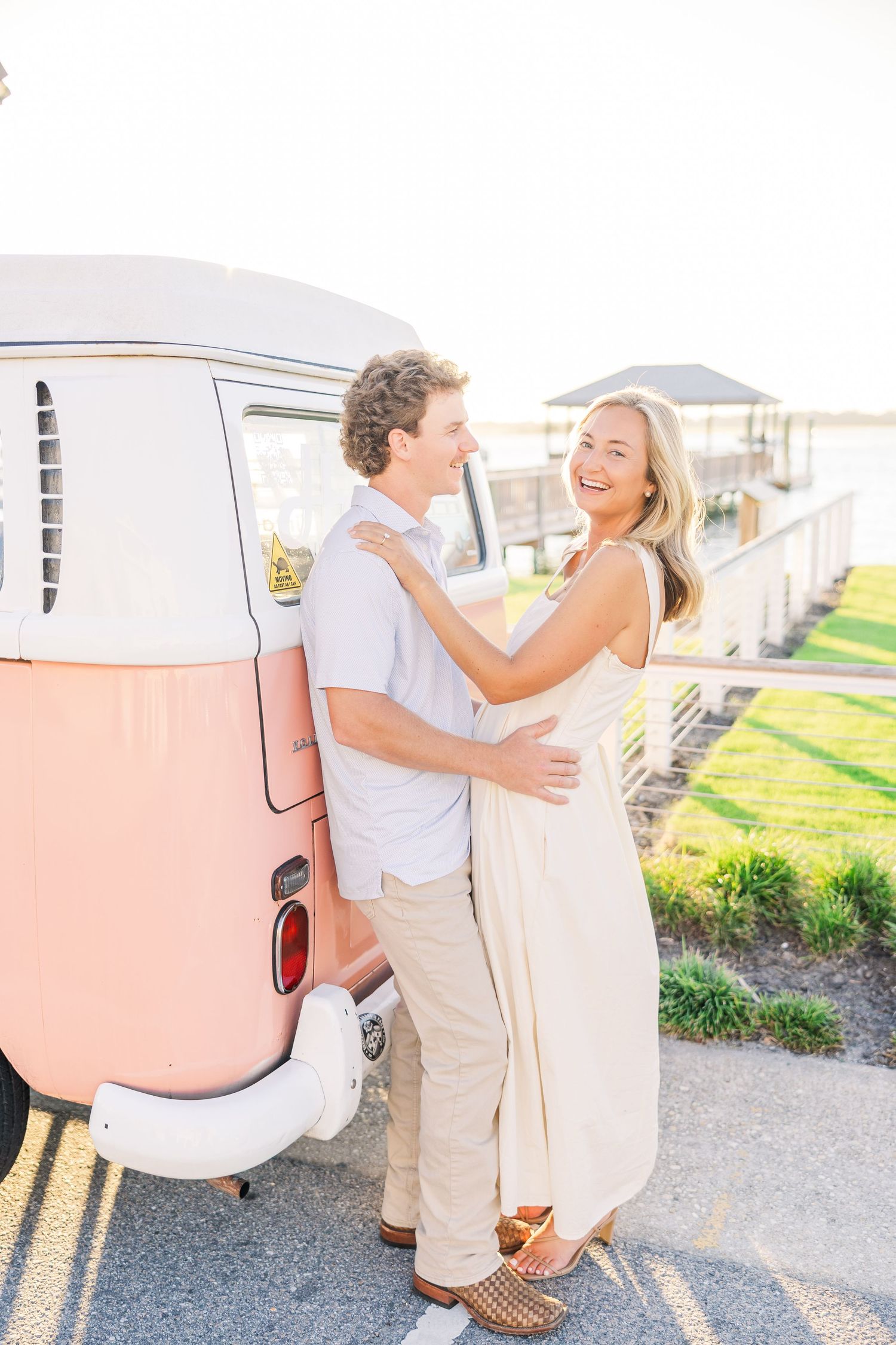 A joyful couple embraces next to a vintage pink and white van on a sunny day.