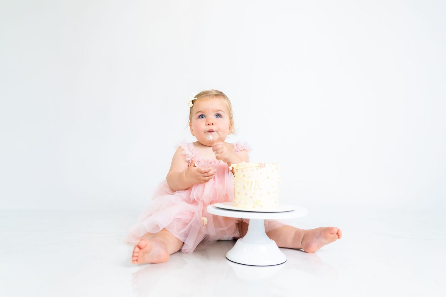 Baby in pink tulle dress crawls near white cake stand during photo shoot.