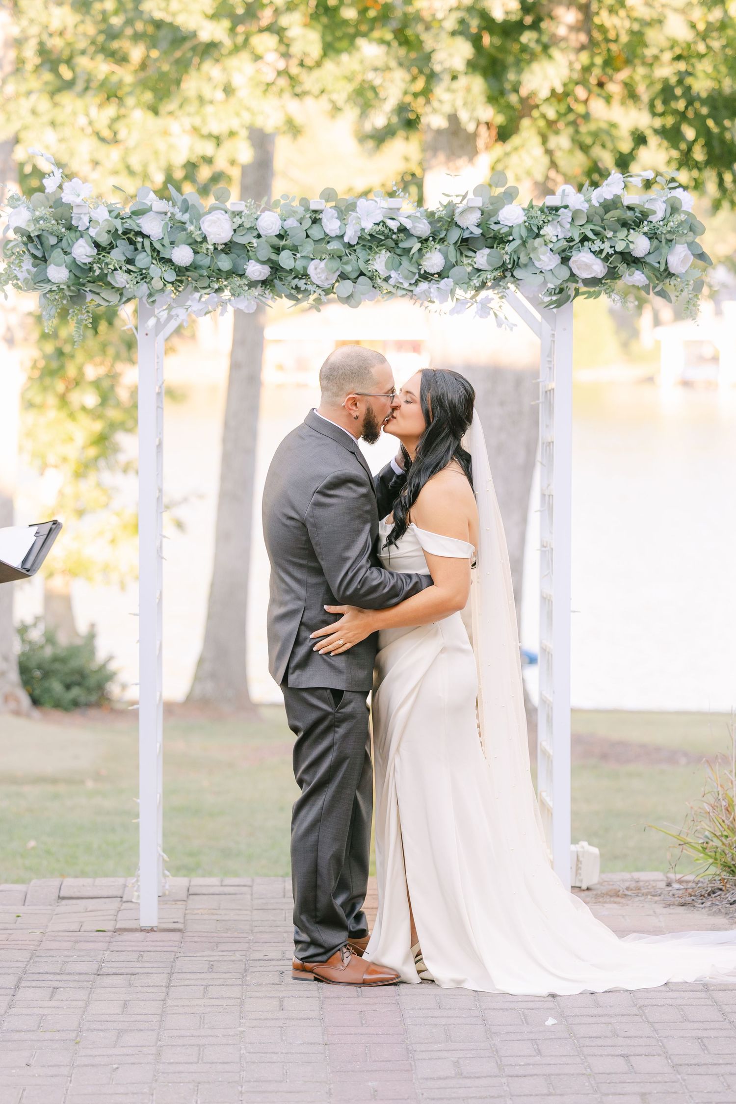 Newlyweds sharing first kiss under floral arch at outdoor wedding venue wedding groom bride lake Gaston photographer light airy