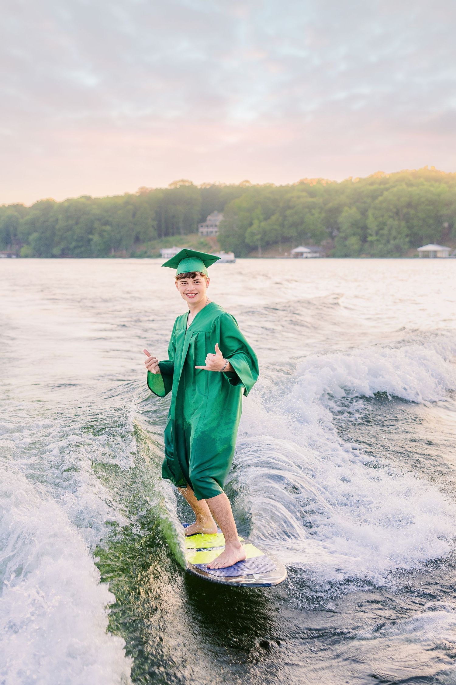 Graduate in green cap and gown surfing on a lake, giving thumbs up, celebrating graduation outdoors.