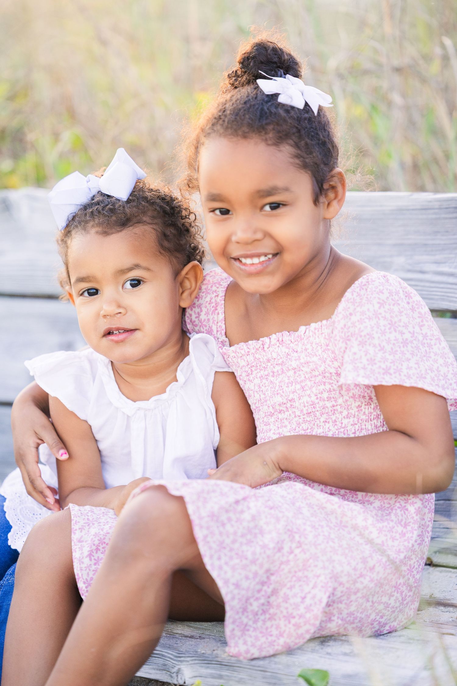 Two children wearing matching hair bows and dresses smile while sitting together on a wooden bench.