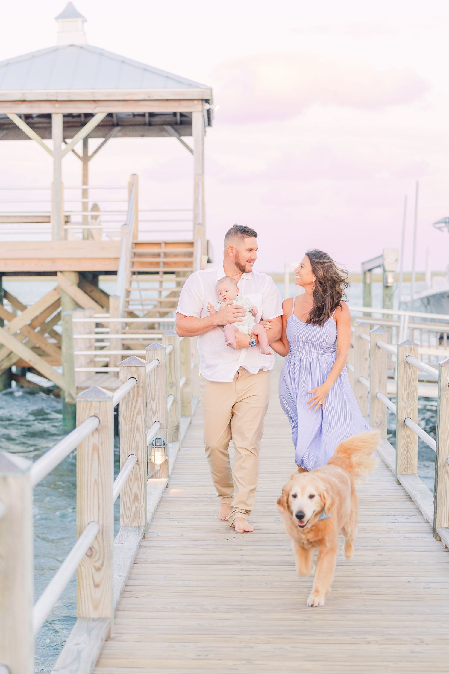 A couple strolls with their golden retriever on a waterfront pier at sunset.