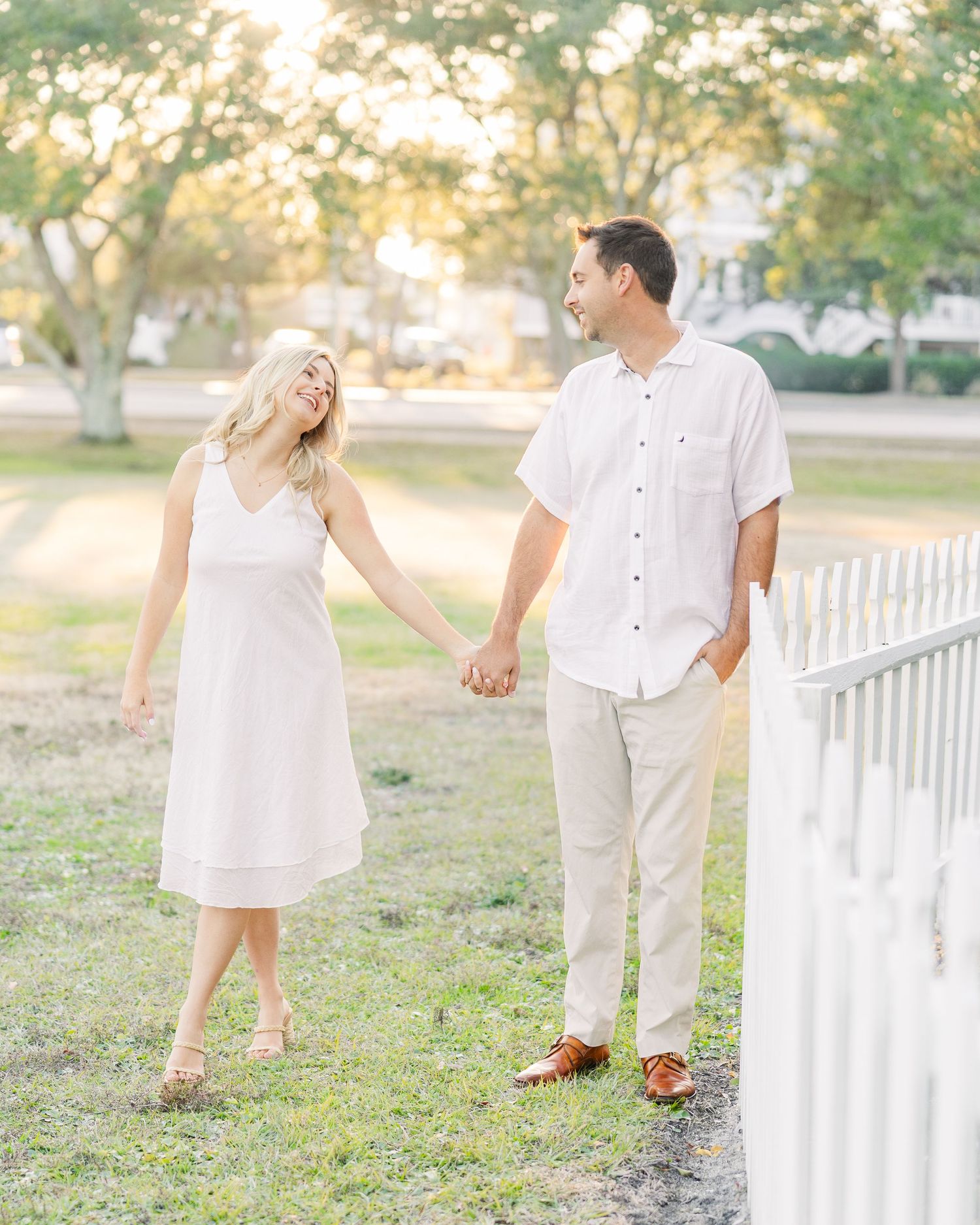 Two people in white outfits pose playfully near a white picket fence outdoors.