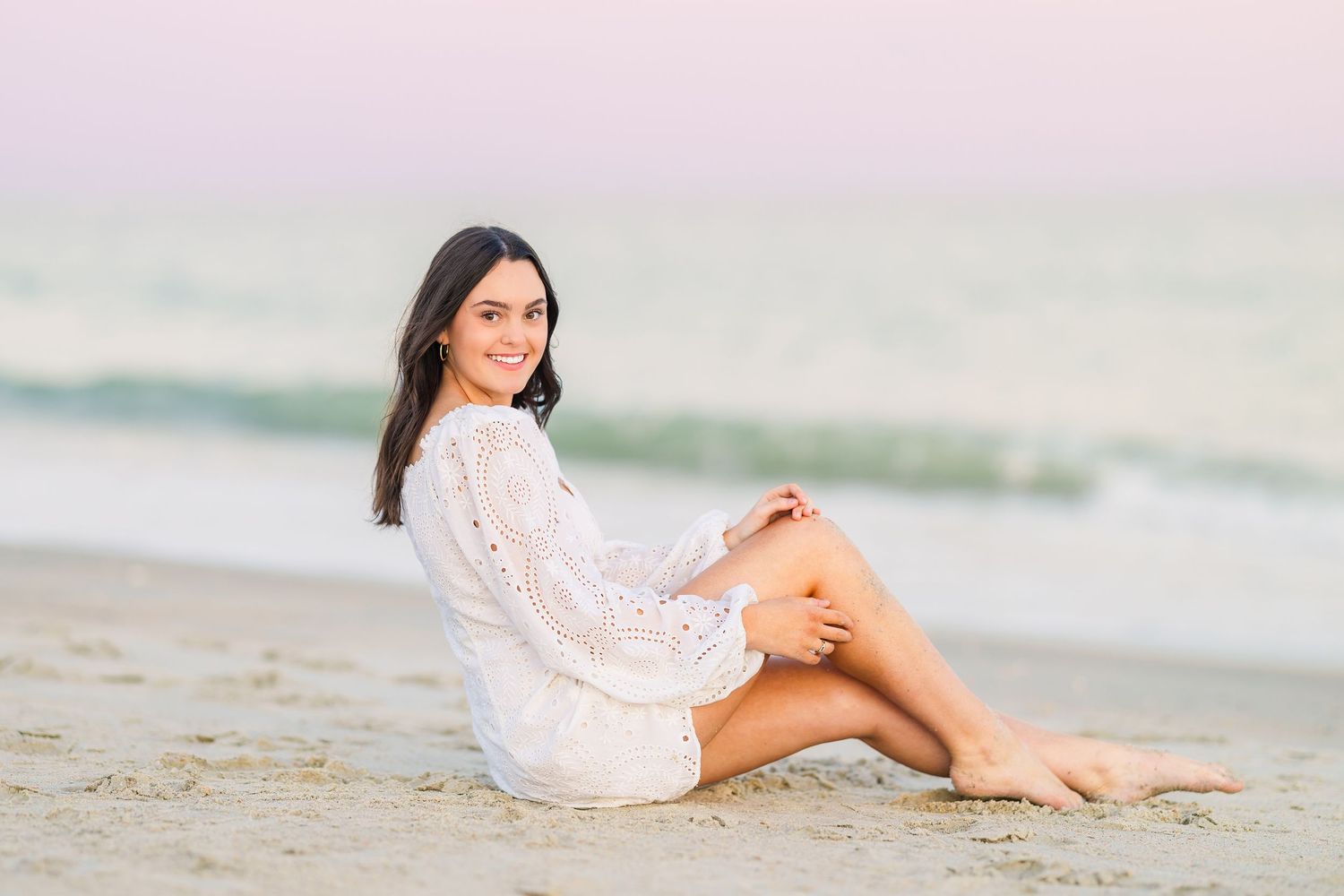 Relaxed beach pose on sand wearing white lace dress during golden hour.