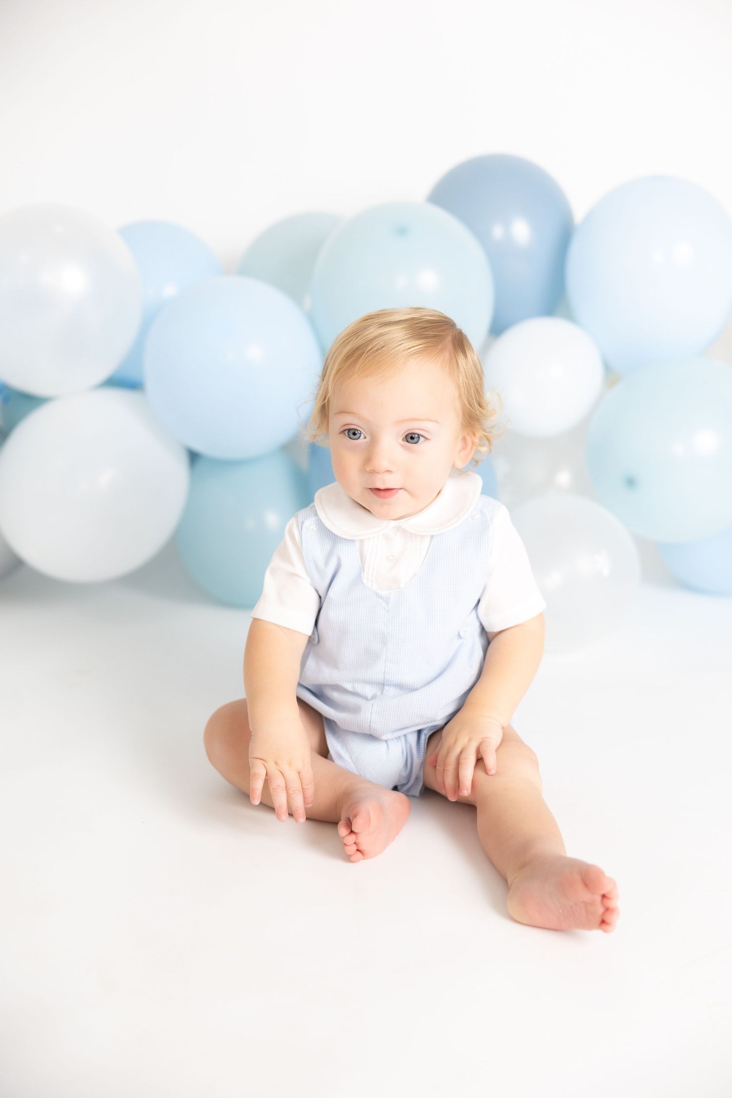 Baby in white outfit crawls on white floor with blue and white balloon garland backdrop.