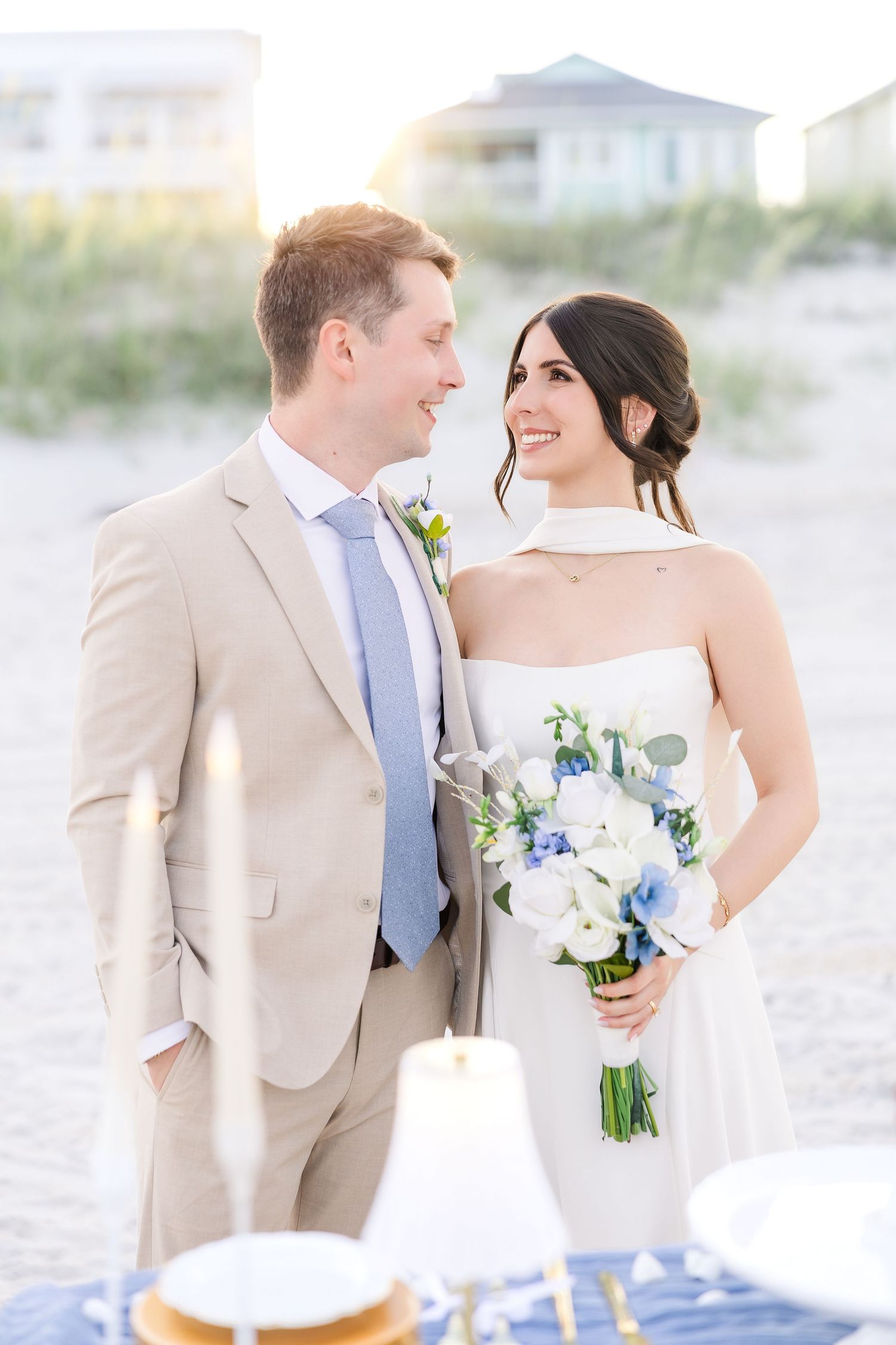 Bride and groom share a romantic glance on the beach, she holds a blue and white bouquet at sunset in Carolina Beach near Wilmington, NC.