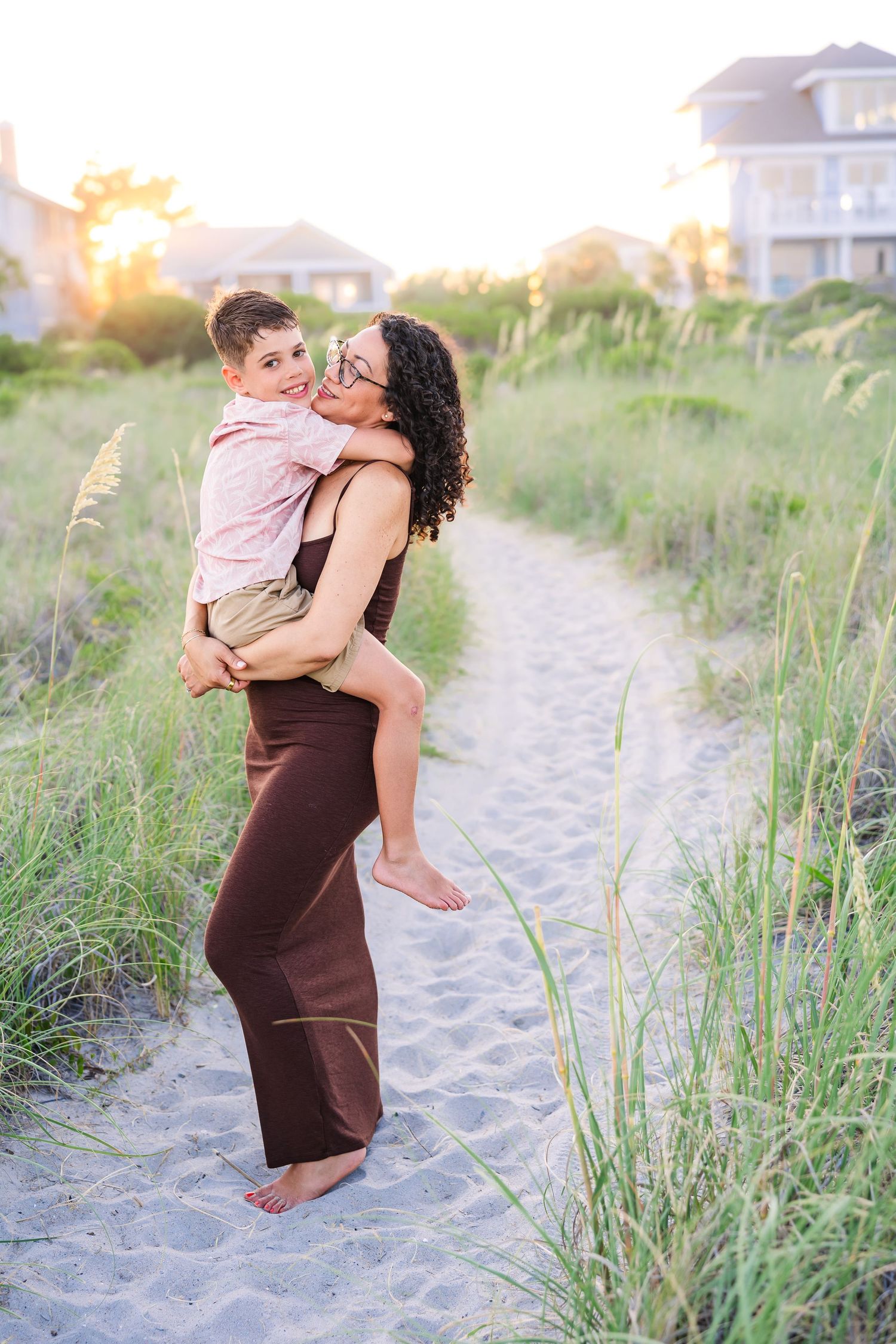 A mother holds her young son on a sandy beach path surrounded by sea grass at golden hour.