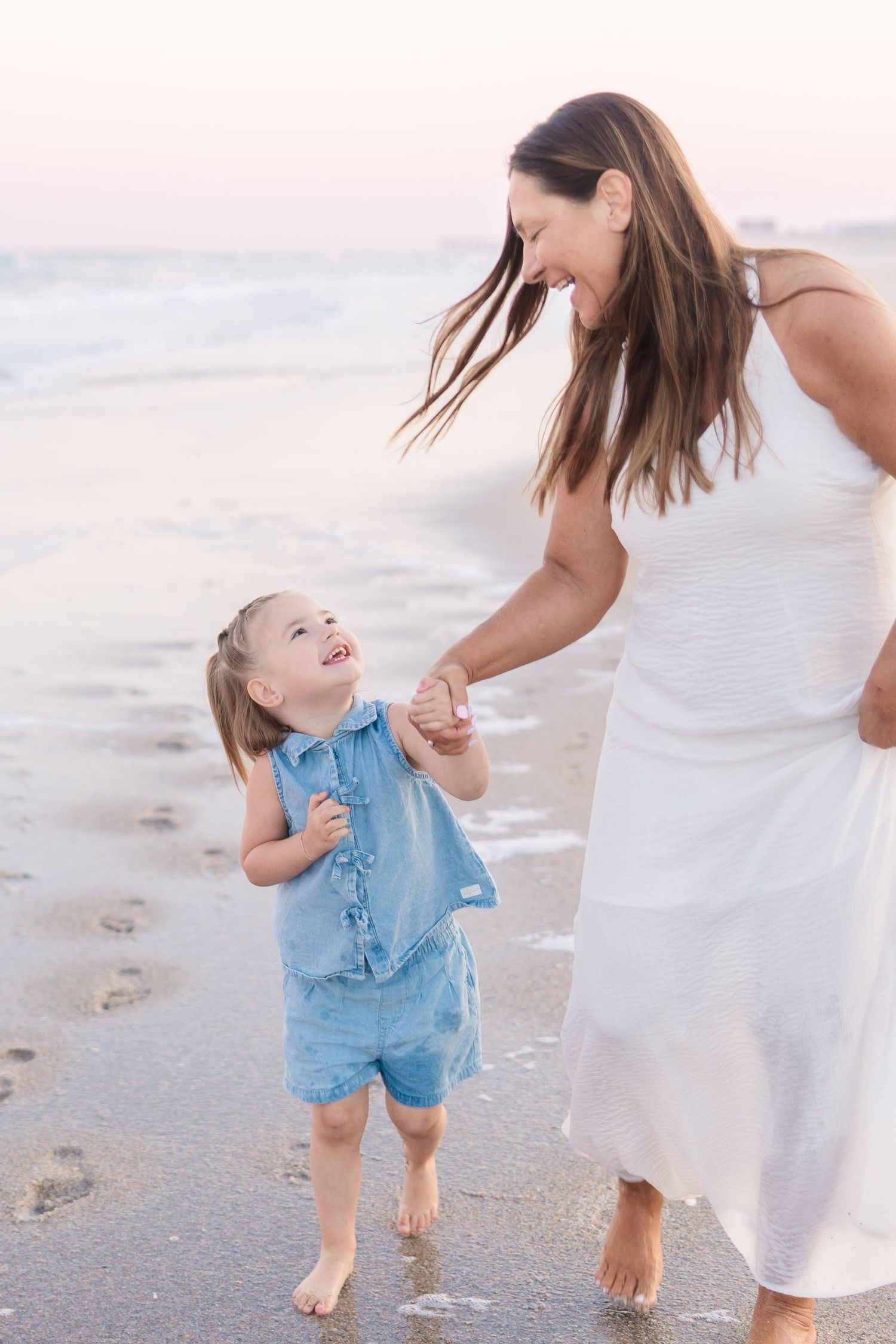 Laughing mother in white dress holds hands with toddler girl in blue romper on ocean shore.