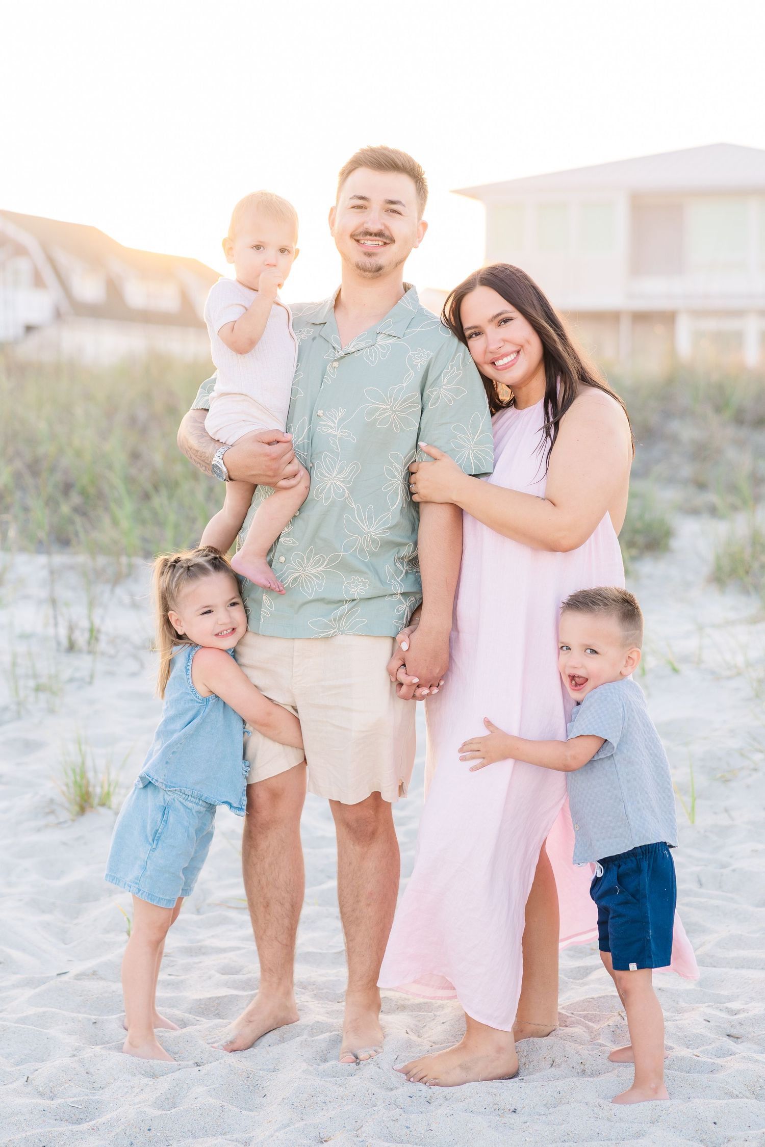Smiling family of five stands on sandy beach, father holding baby while children hug parents.