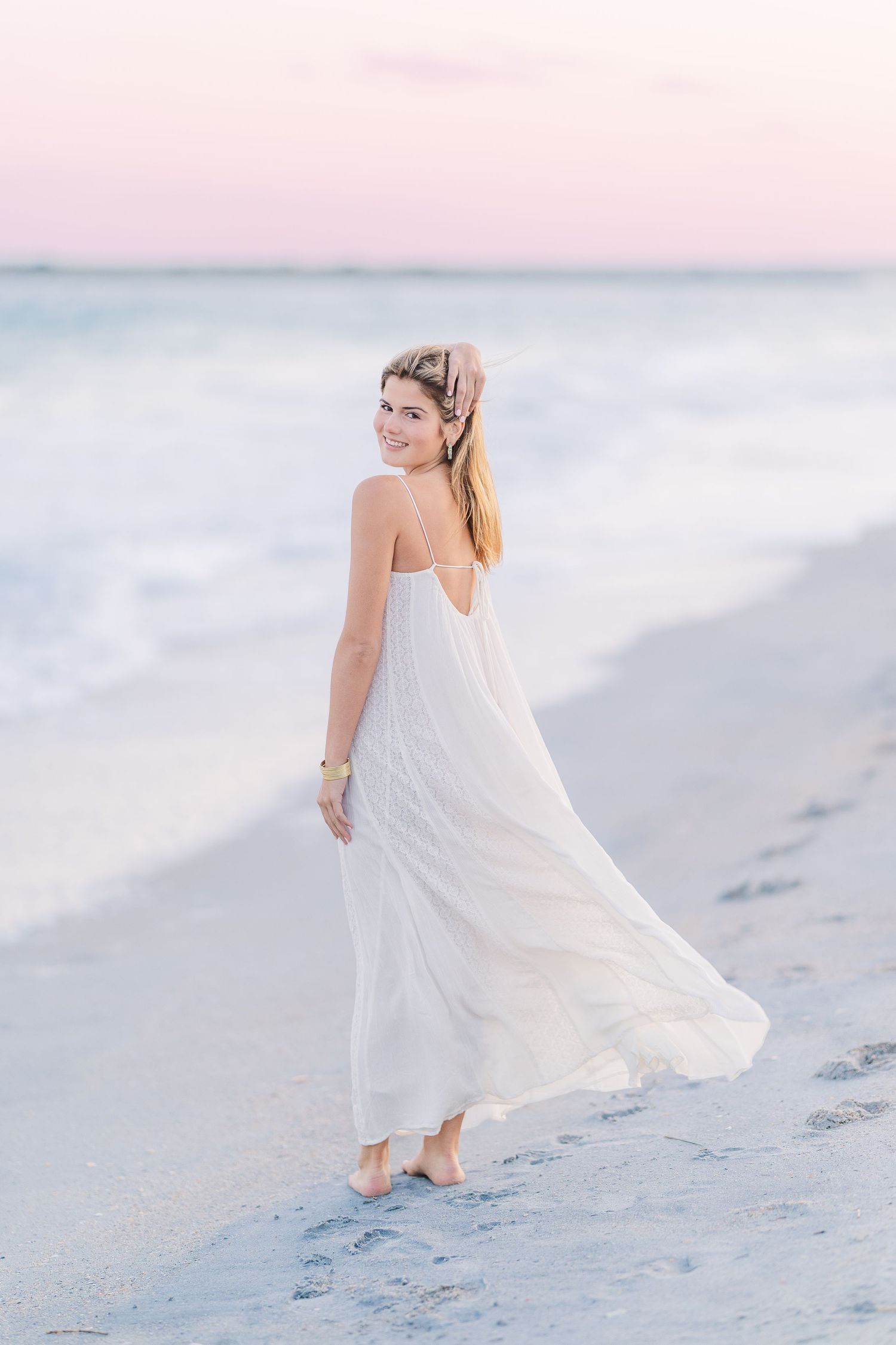 Romantic coastal photoshoot featuring billowing white dress and soft pink sunset hues.