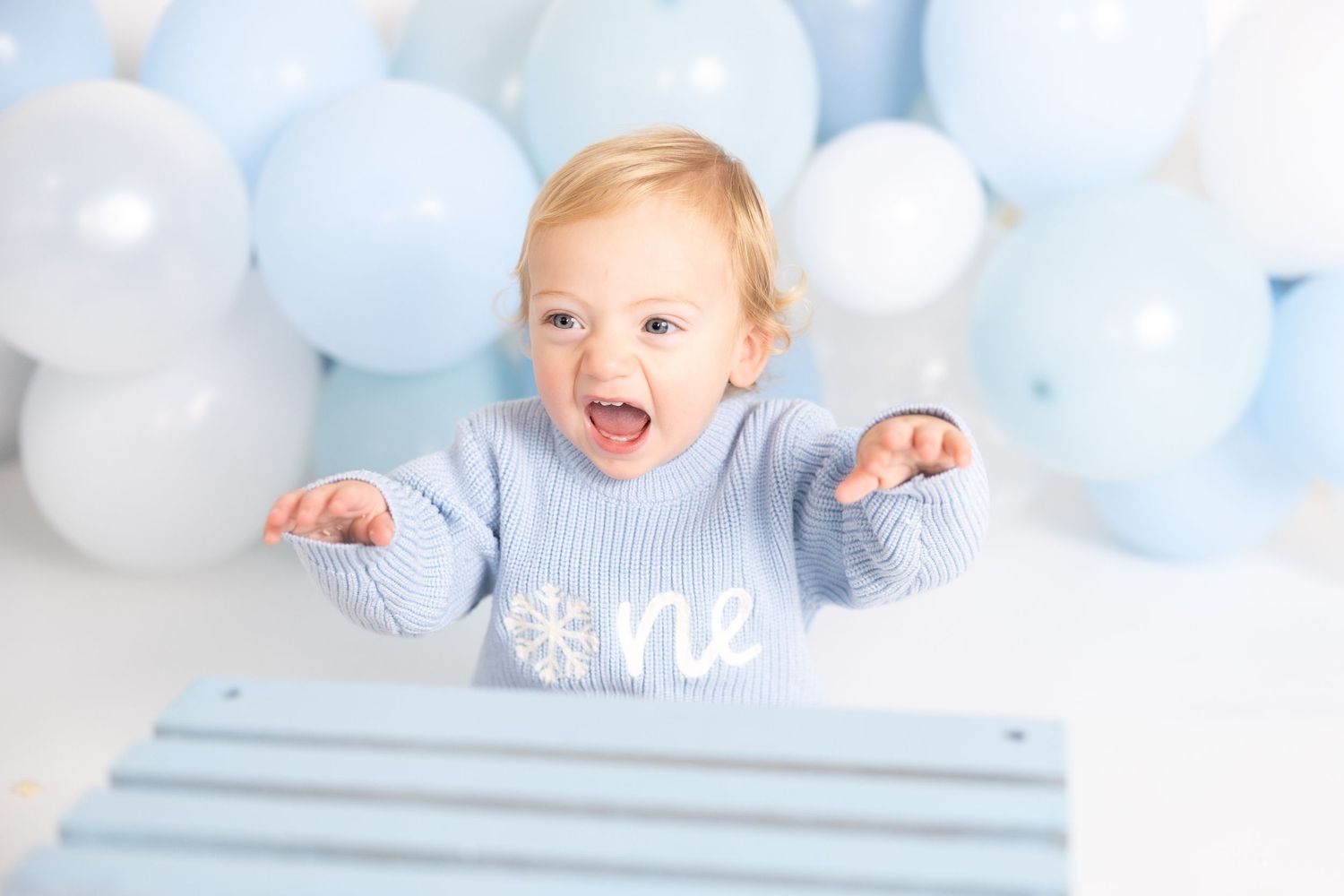 Excited baby in blue sweater plays with wooden crate surrounded by balloons.