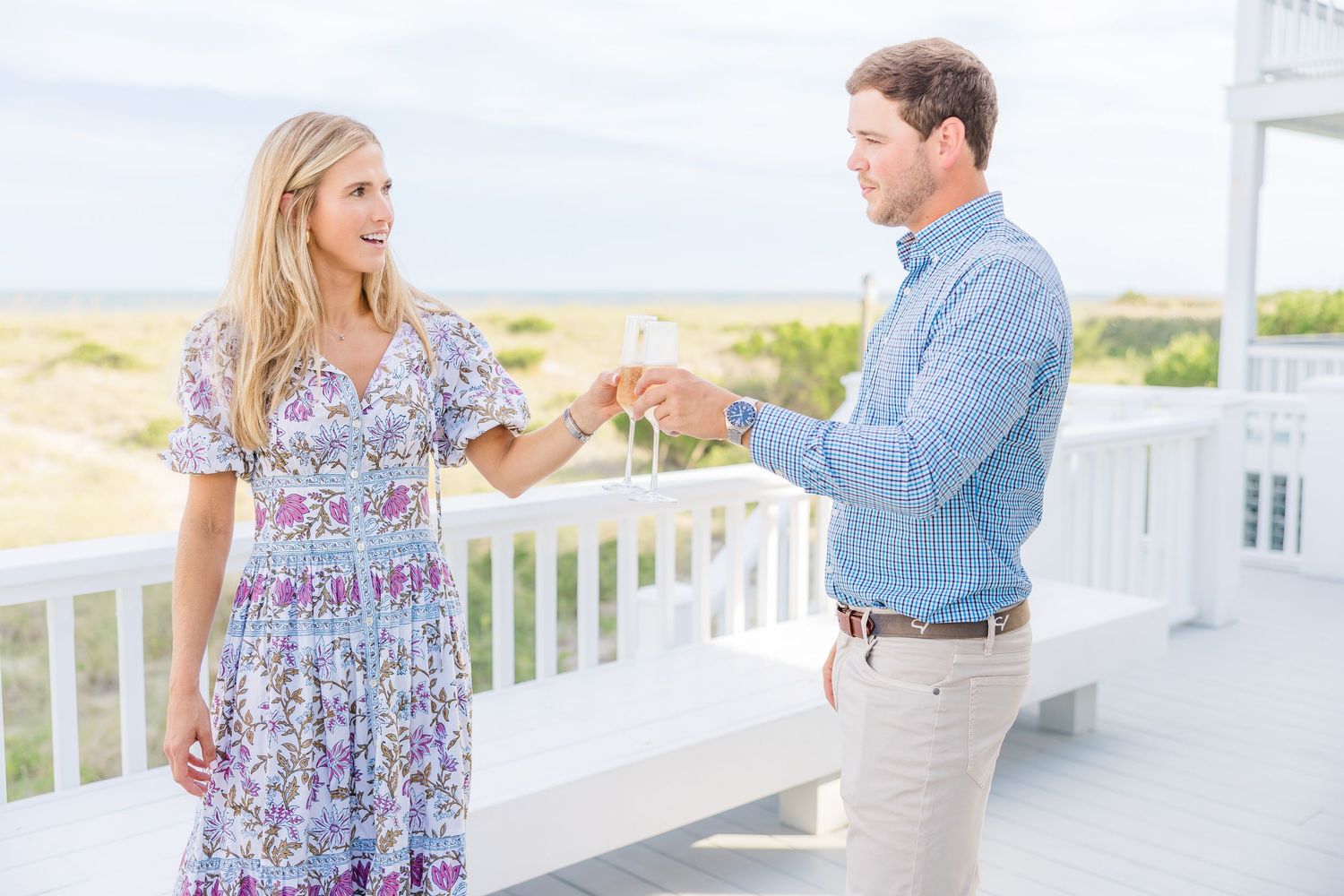 A couple shares a romantic moment on a white wooden deck overlooking the water at sunset.