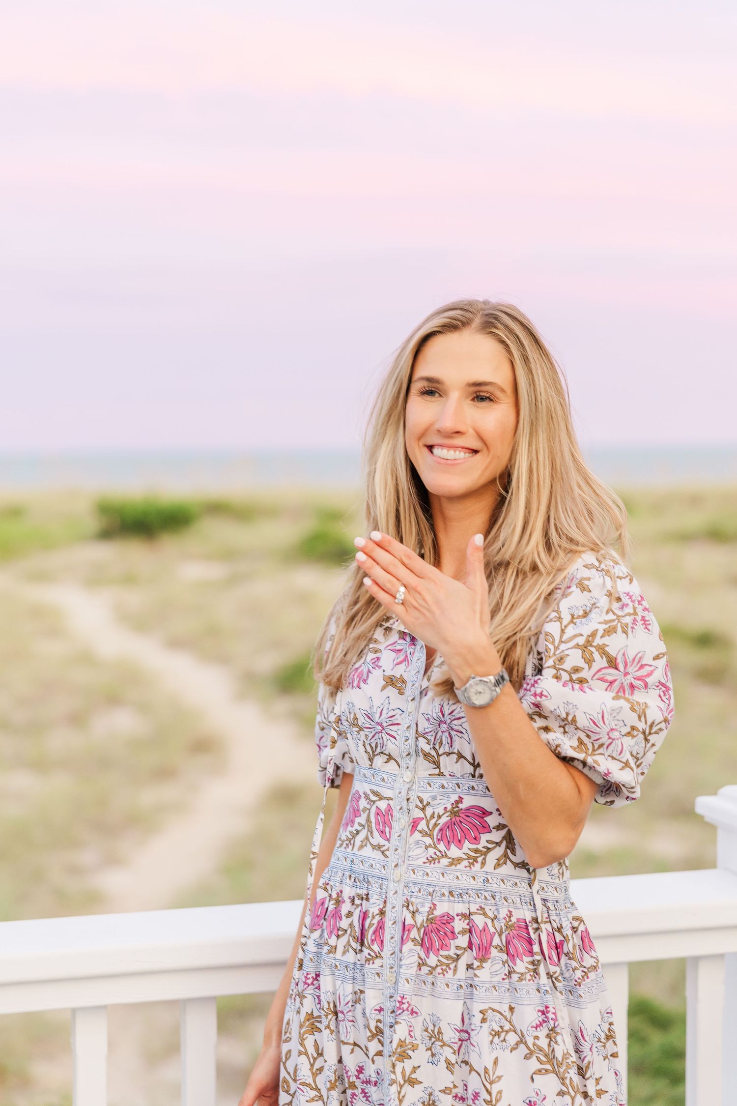 A woman in a floral dress poses on a beach house deck during sunset.