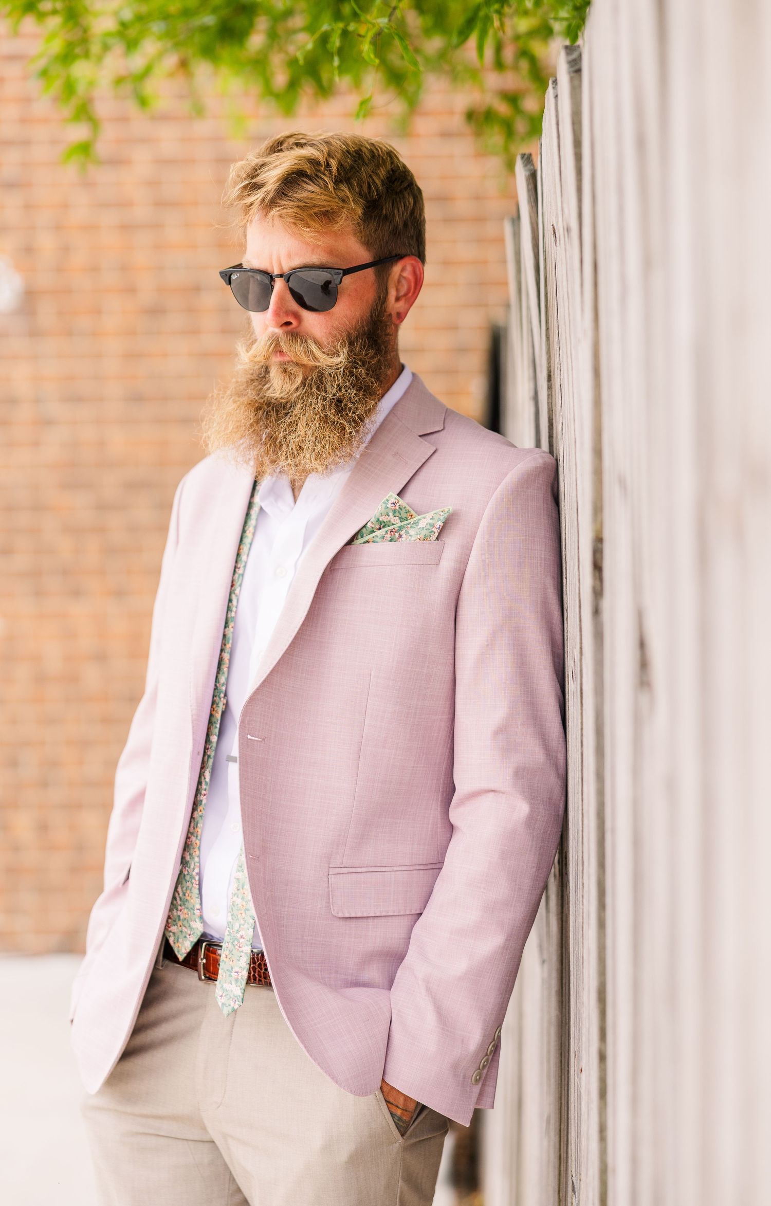 A groom in a light pink blazer and sunglasses poses against a brick wall with green foliage overhead in a backyard wedding in wilmington, nc