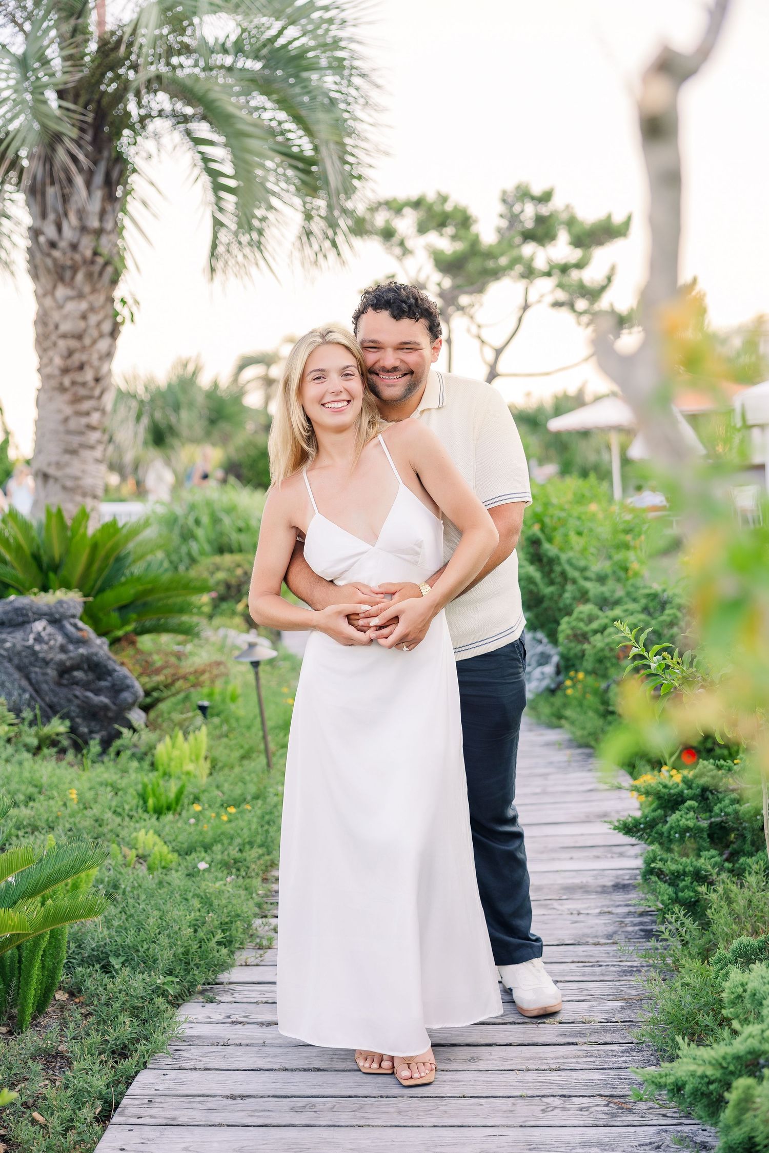 A smiling couple embraces on a wooden boardwalk surrounded by lush tropical greenery, woman in white dress.