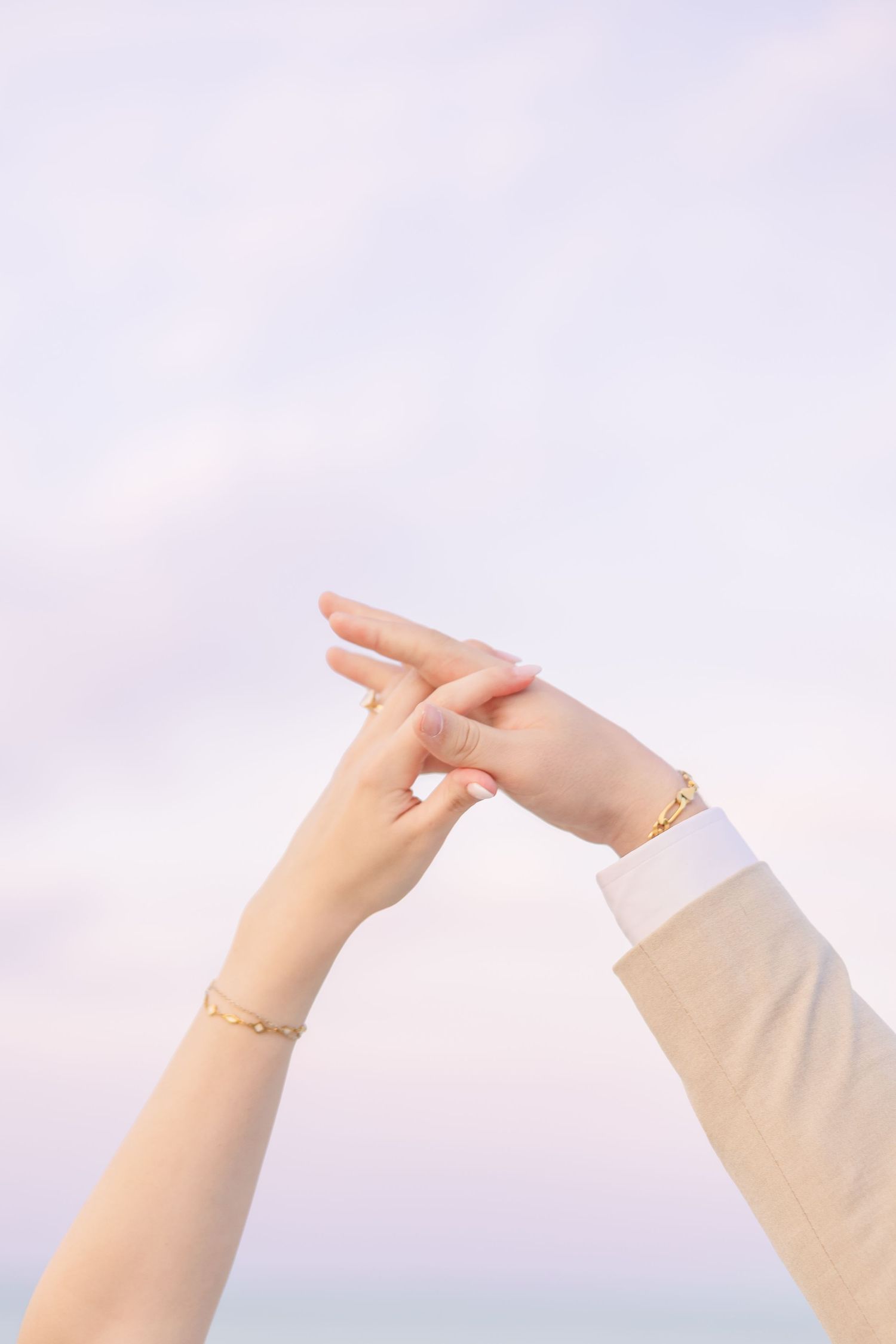 Close-up of bride and groom's hands touching against a soft pastel sky, both wearing gold bracelets in Carolina Beach near Wilmington, NC.