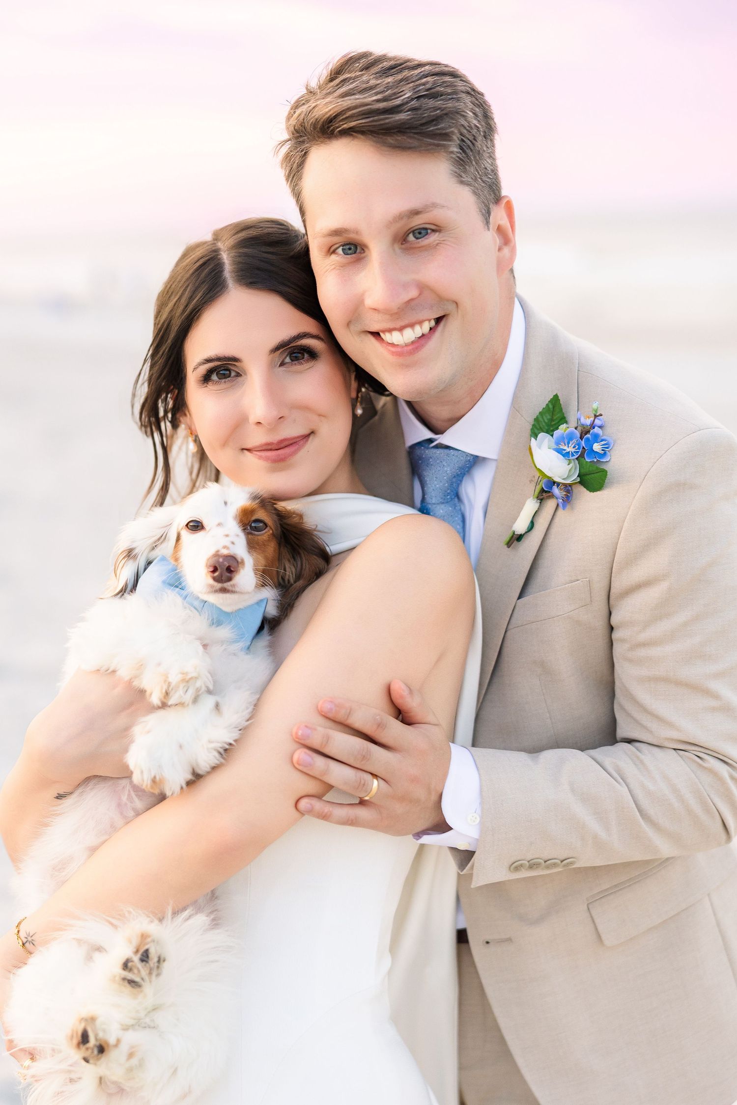 Happy newlyweds pose with their adorable dog on the beach, groom in tan suit with blue tie in Carolina Beach near Wilmington, NC.