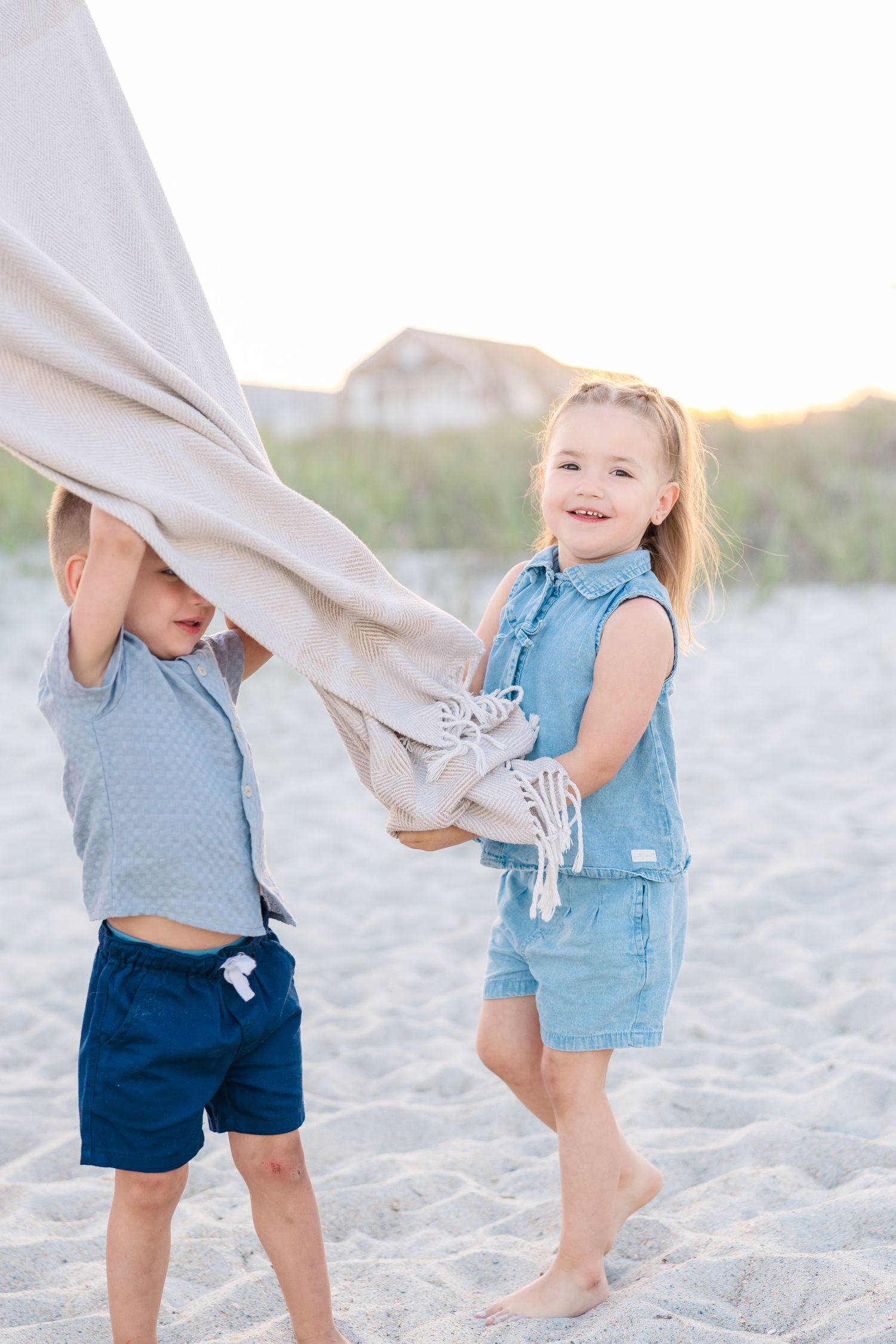 Two young siblings play with a blanket on a sandy beach at sunset, girl in denim smiling brightly.