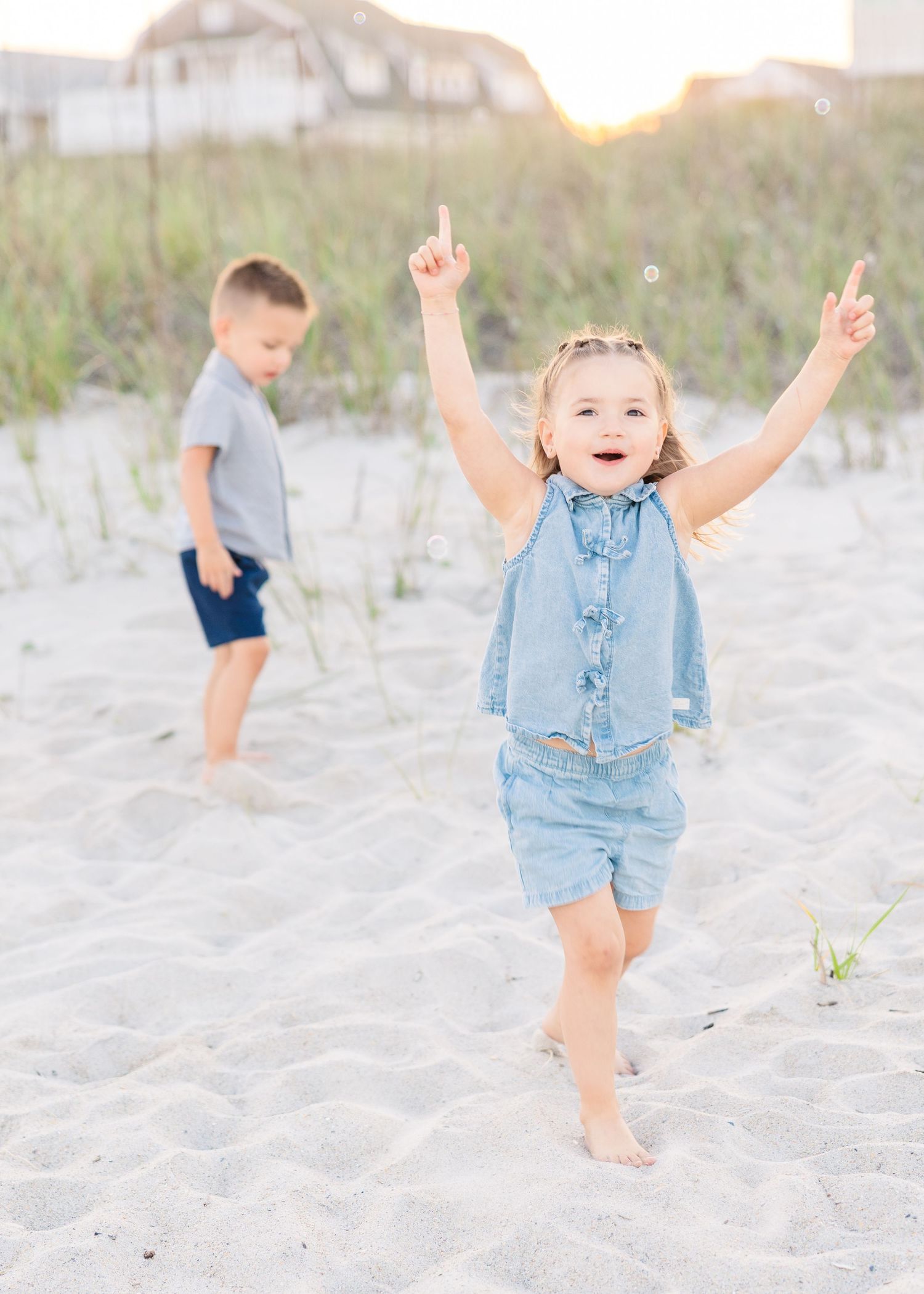 Joyful little girl in denim outfit raises arms on sandy beach while boy stands in background.