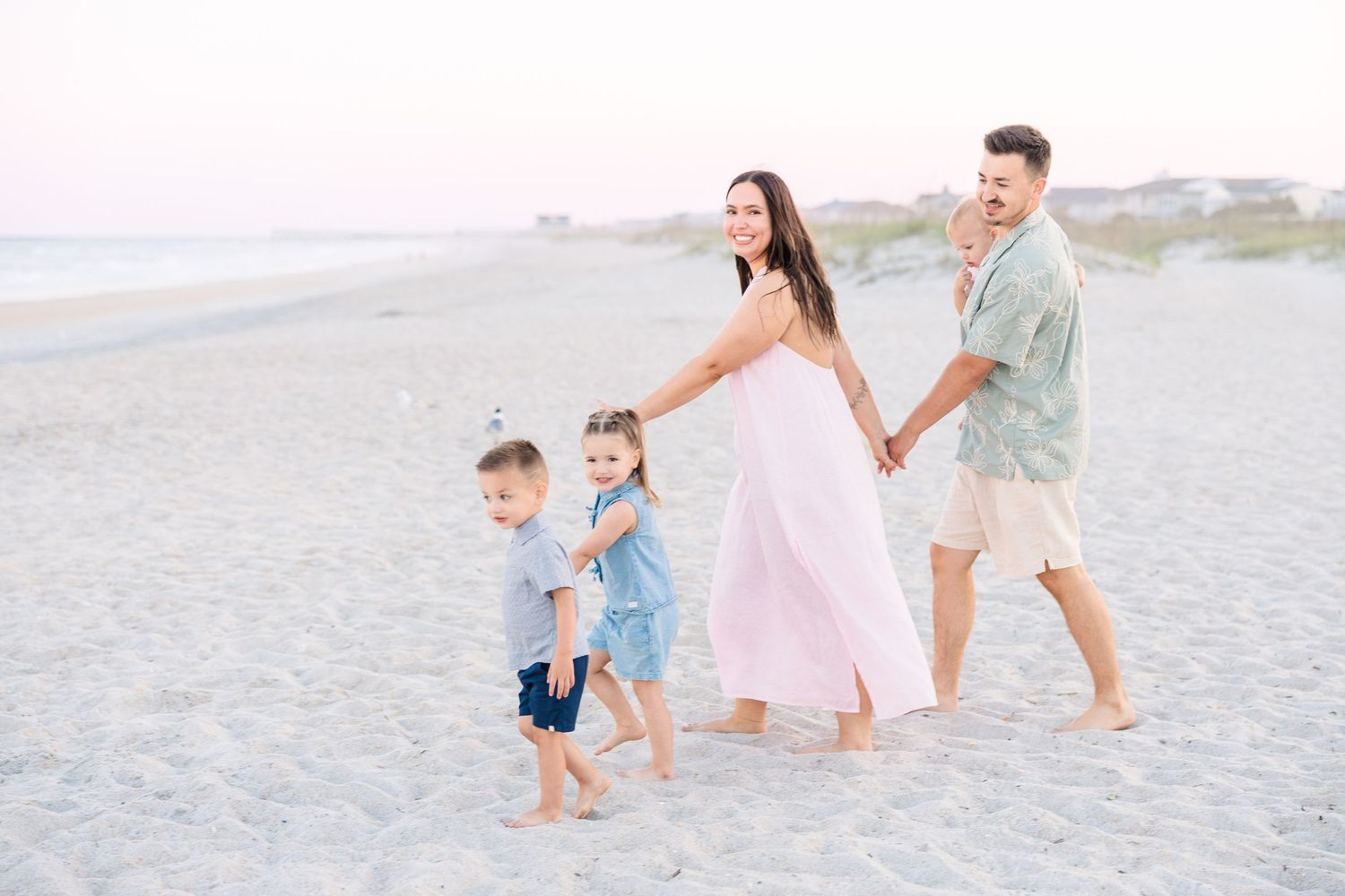 Family of four walking hand-in-hand on a sandy beach at sunset, parents and two young children in pastel outfits.