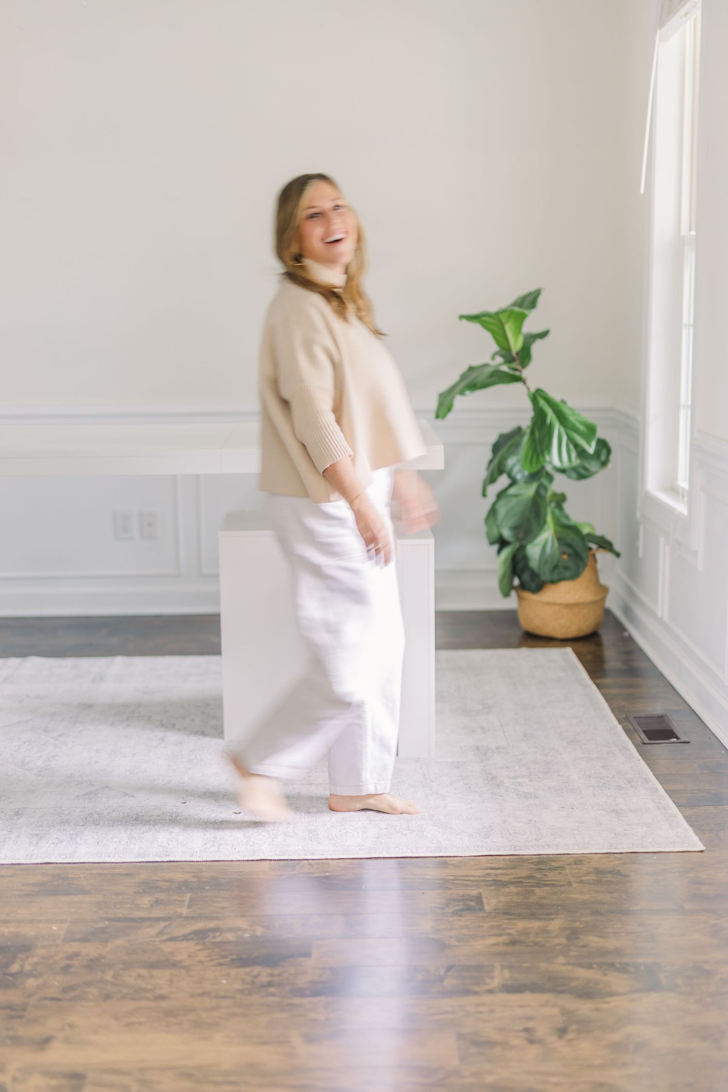 Candid lifestyle photo of someone in cream sweater and white pants standing by houseplant.