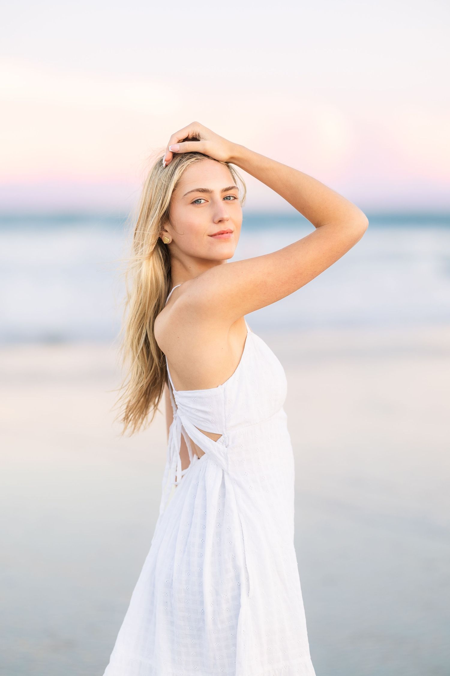 Woman in flowing white dress poses elegantly on beach at sunset.