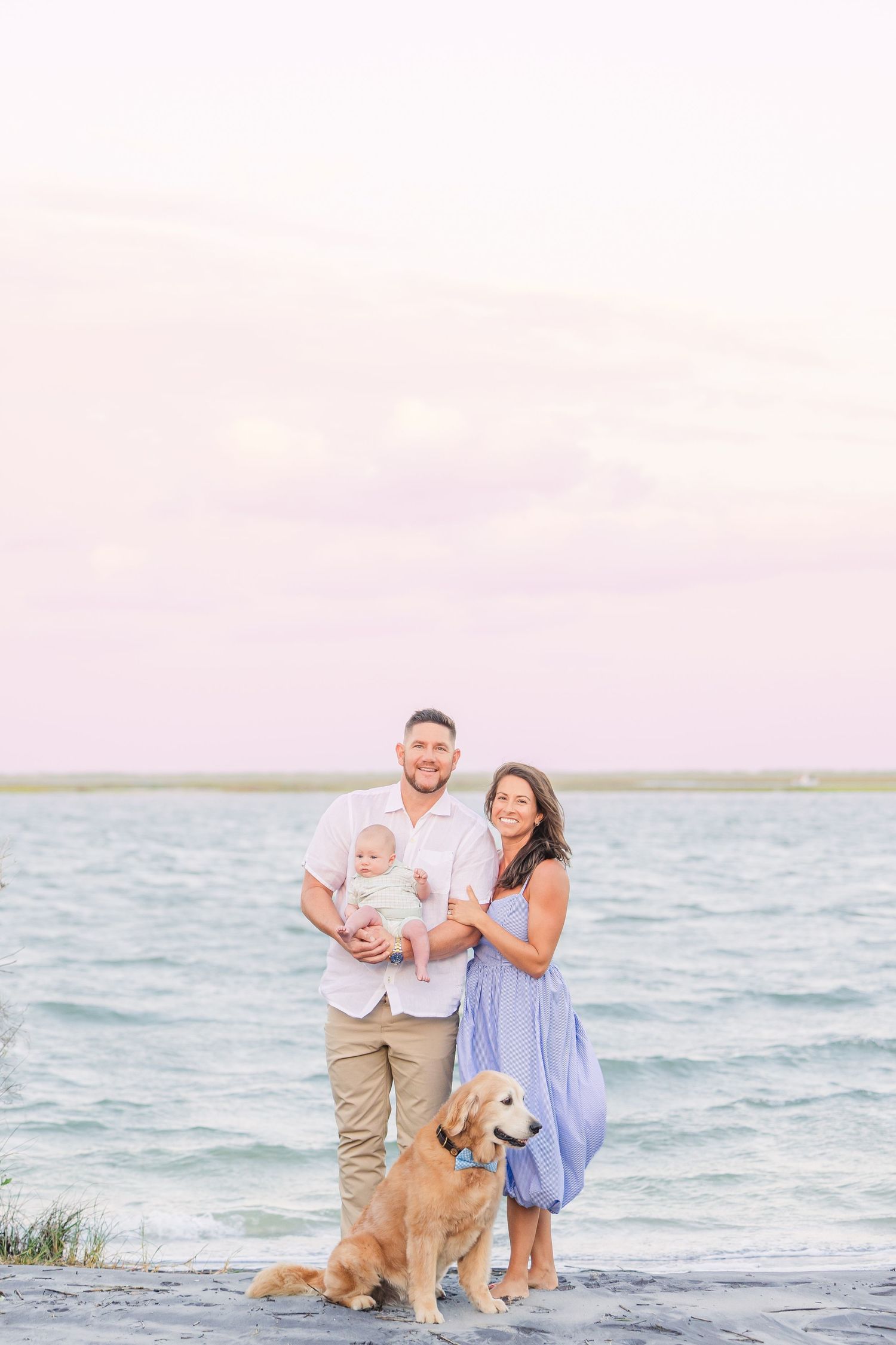 A couple and their golden retriever pose for photos at the beach during sunset.