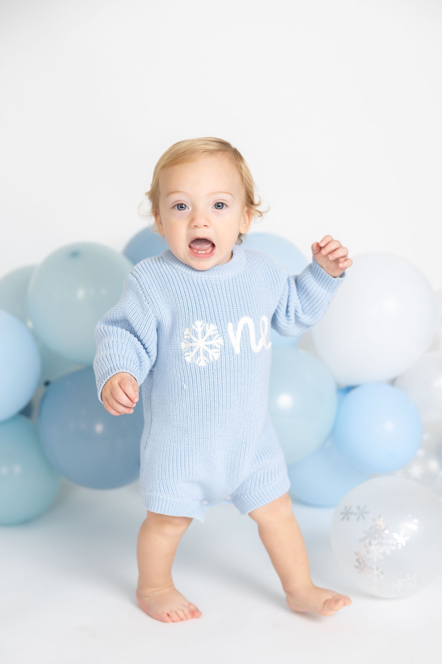 Baby in ribbed blue sweater plays near festive balloon arrangement on white background.