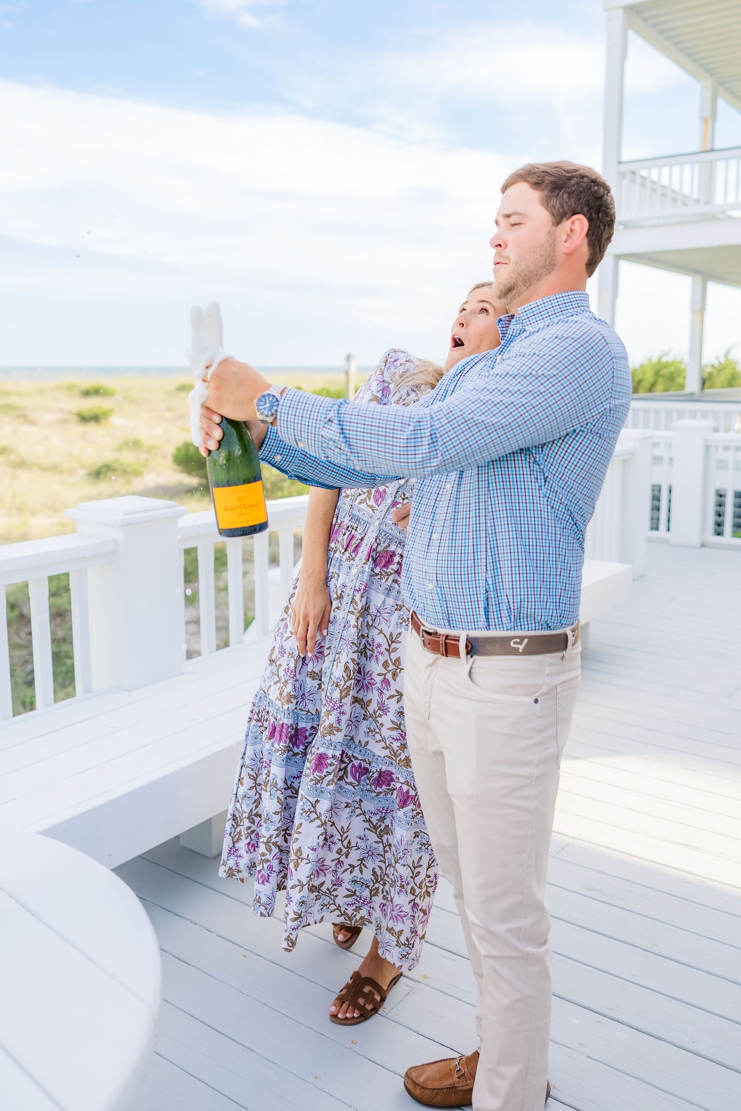 A couple opens champagne together on a white beachfront deck.