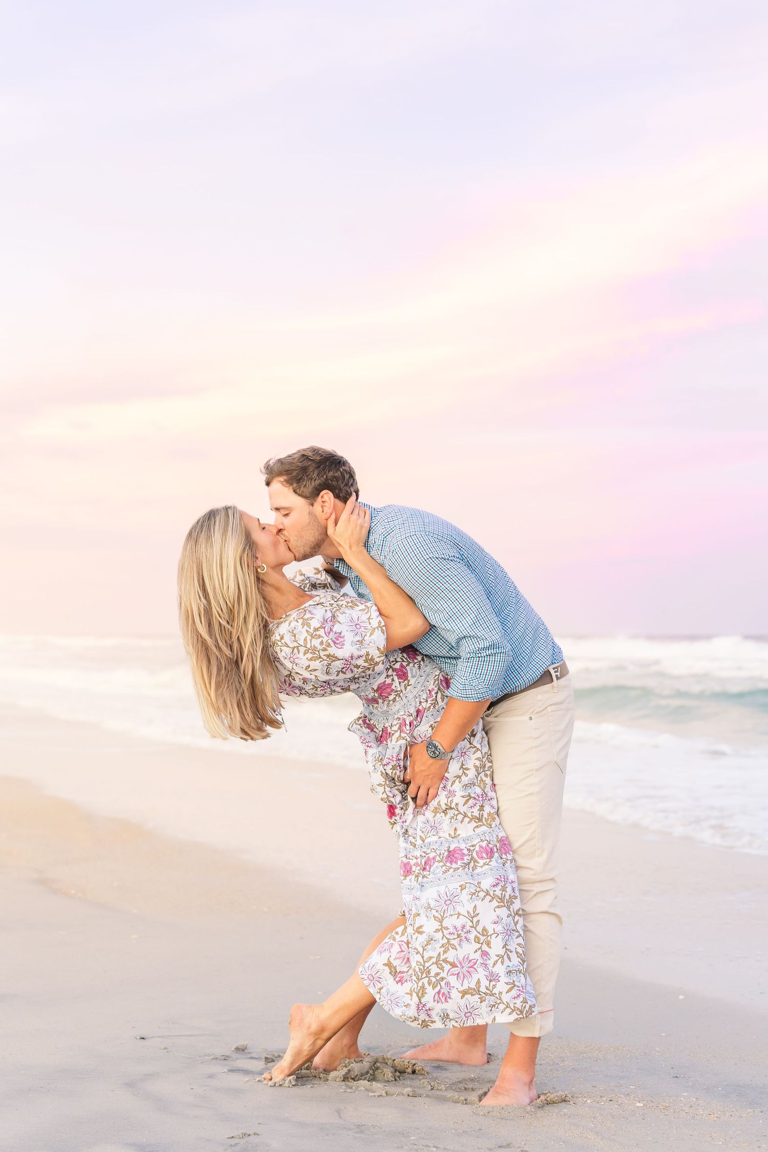 Couple sharing romantic dip kiss on beach with pink sunset sky.