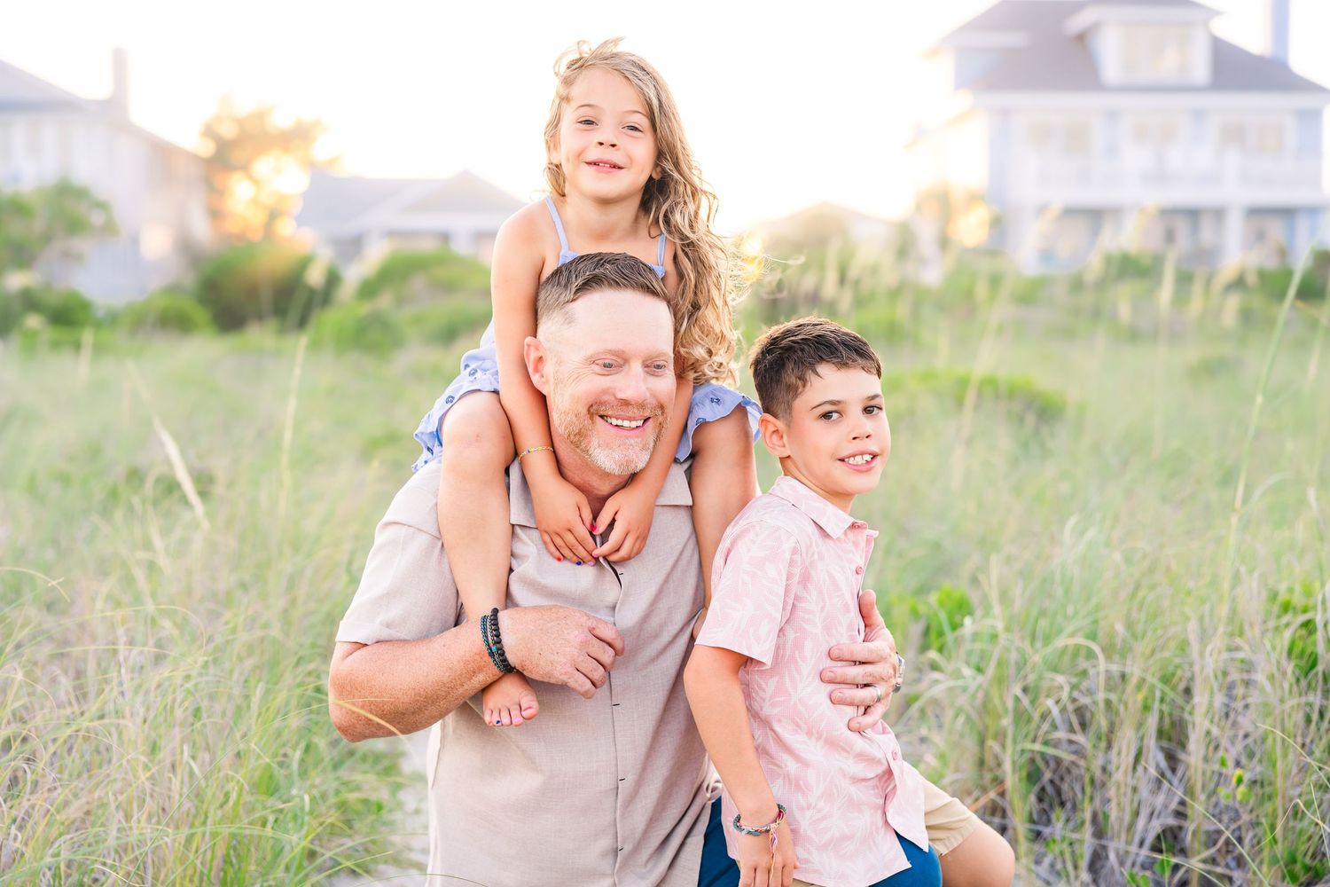 A man smiles with a girl on his shoulders and a boy beside him in a sunny beach grass field.