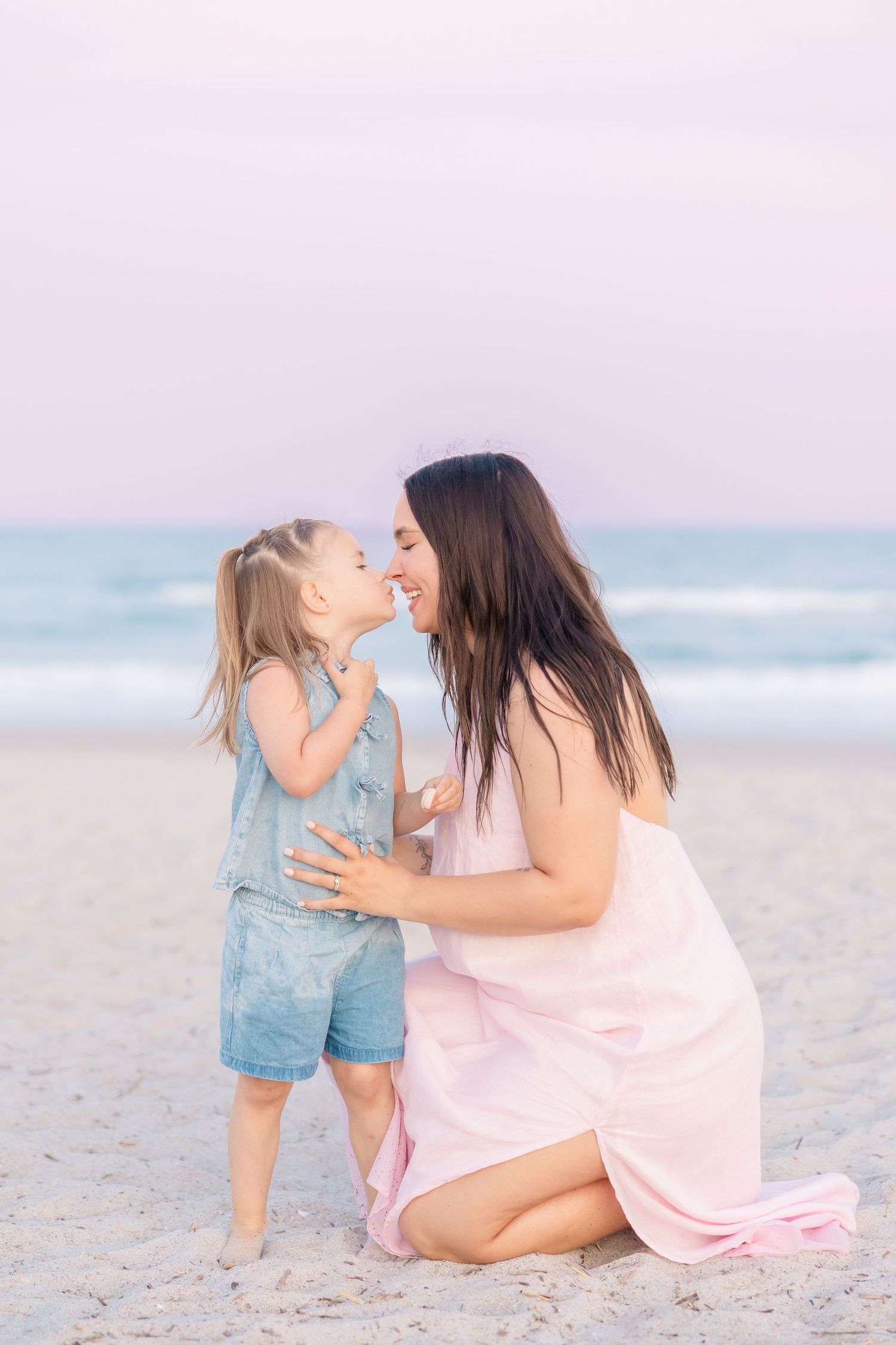 Mother kneels on beach sharing a tender nose-to-nose moment with her young blonde daughter.