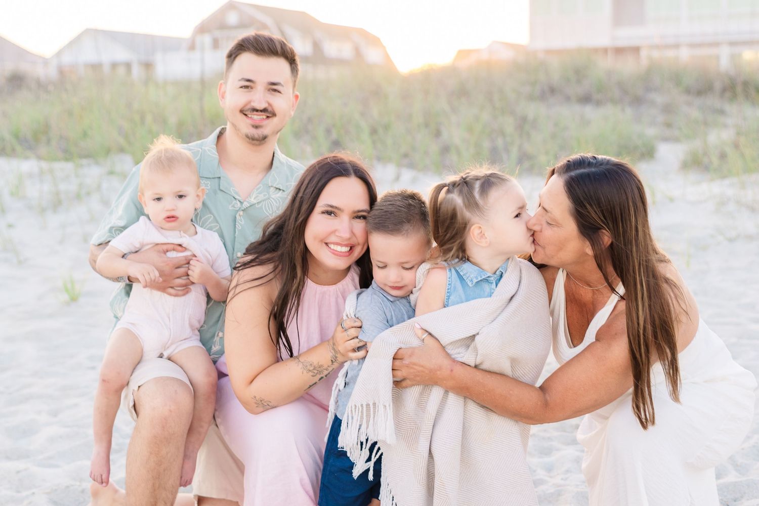 Happy family of six posing together on a sandy beach at golden hour, smiling and laughing near coastal dunes.