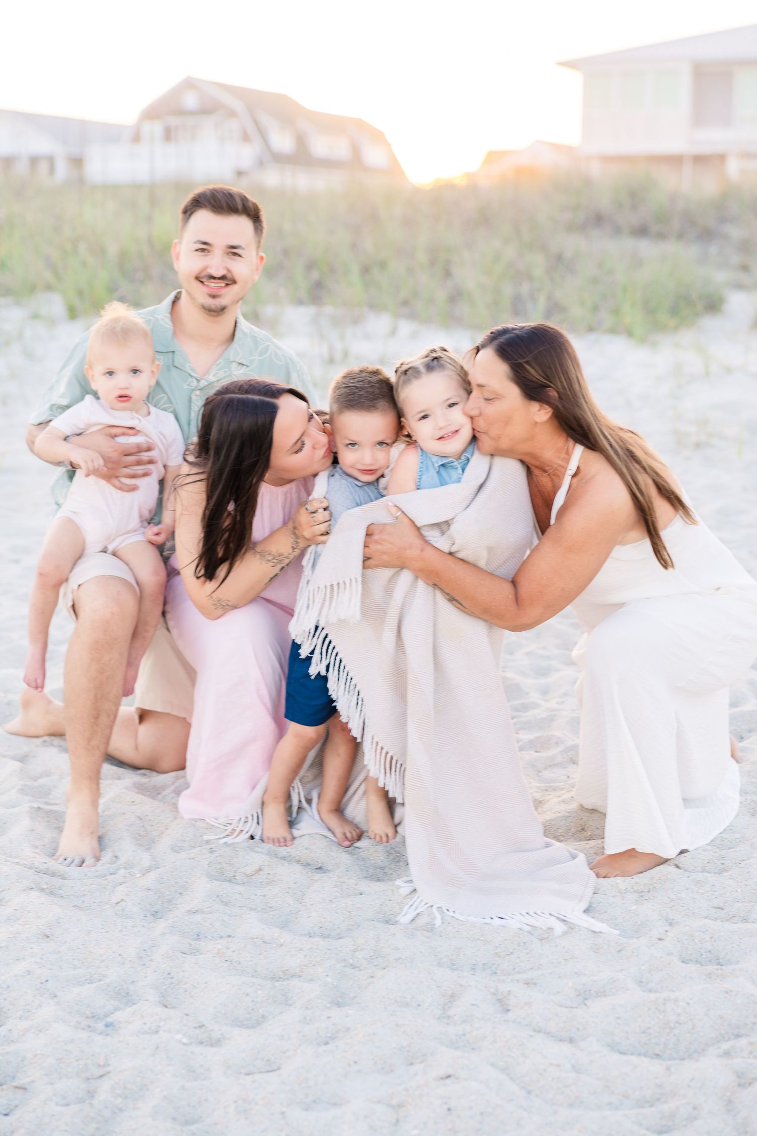Family of six poses together on sandy beach at sunset, with two women kissing a toddler while man holds baby.