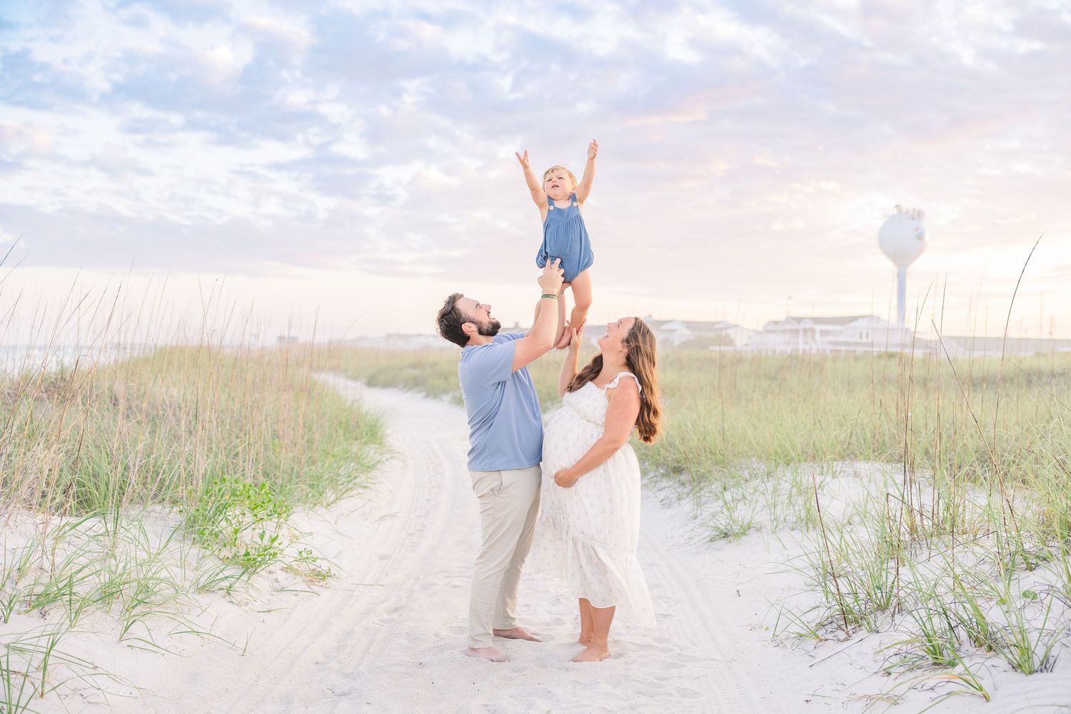 A joyful family on a sandy beach path, dad lifting toddler high in the air while mom watches at sunset.