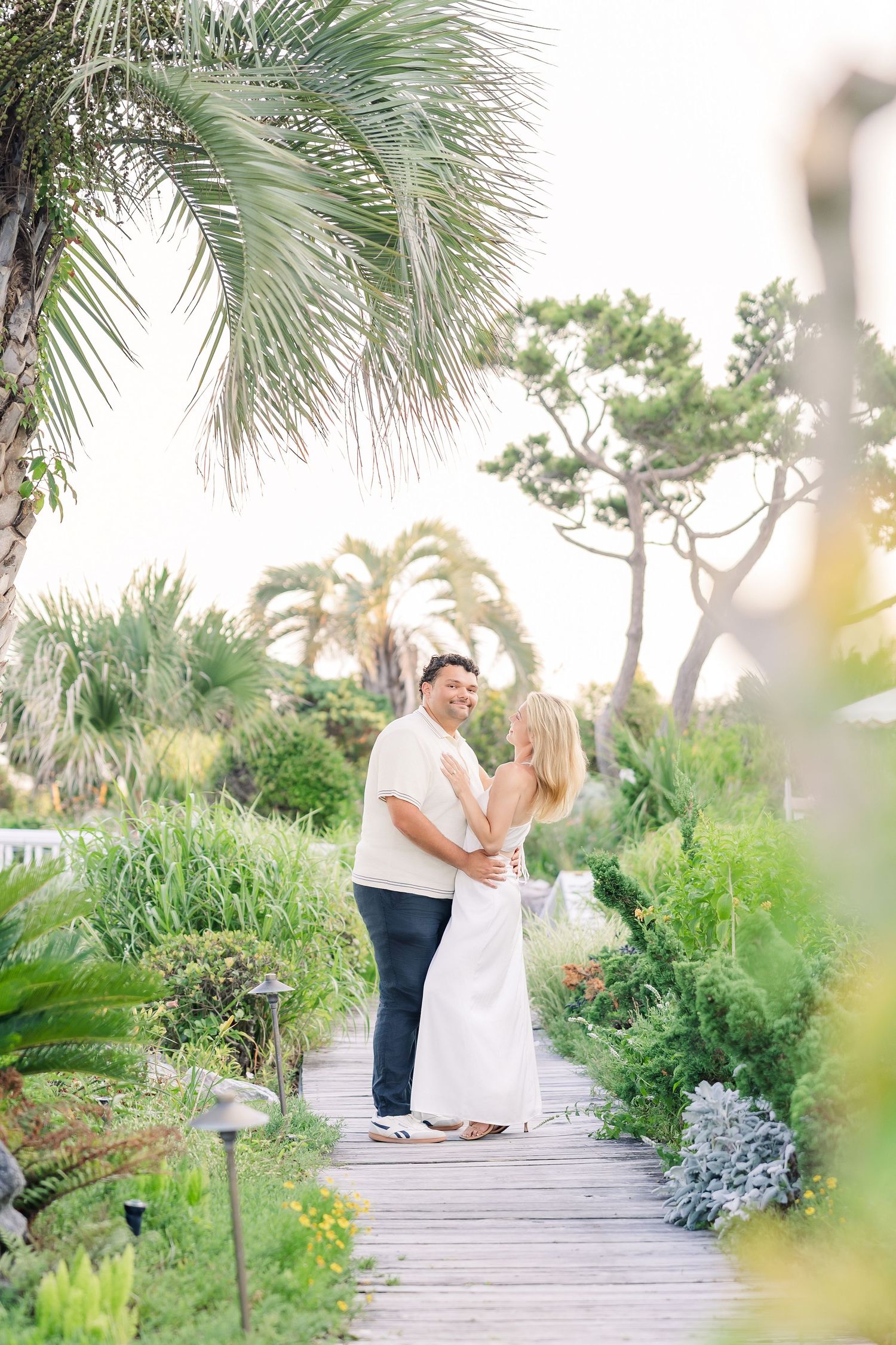 Couple holding hands and smiling on a wooden boardwalk surrounded by lush tropical garden greenery.