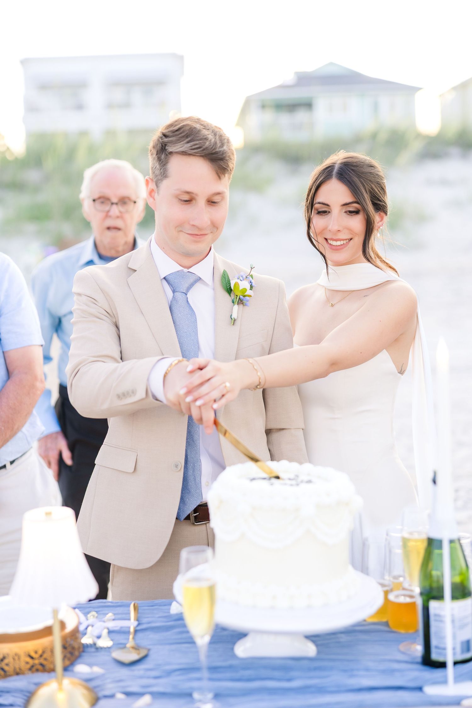 Bride and groom cutting white wedding cake on beach with champagne glasses nearby in Carolina Beach near Wilmington, NC.