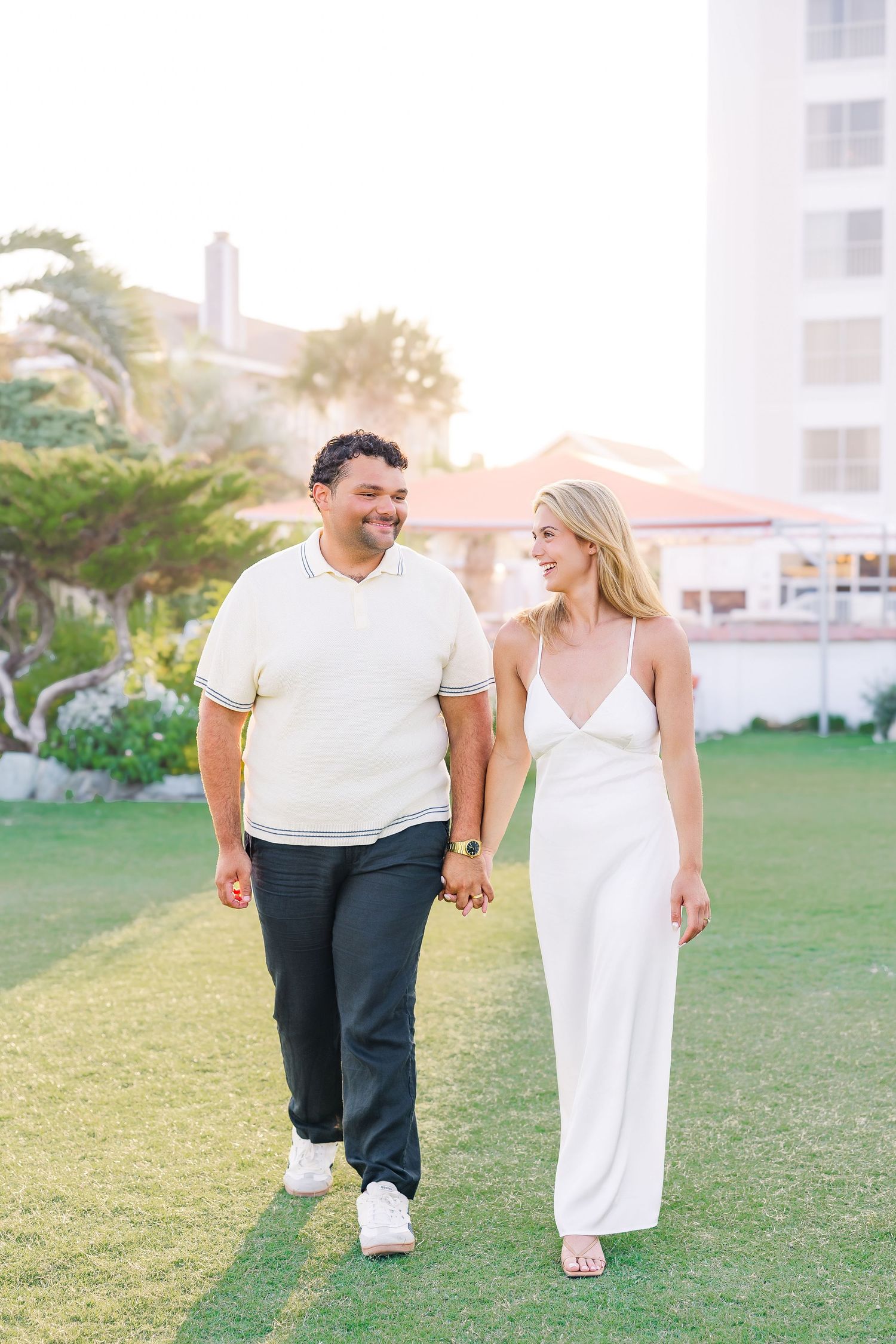 Smiling couple holding hands and walking on green lawn at sunset, woman in white slip dress and man in cream polo.