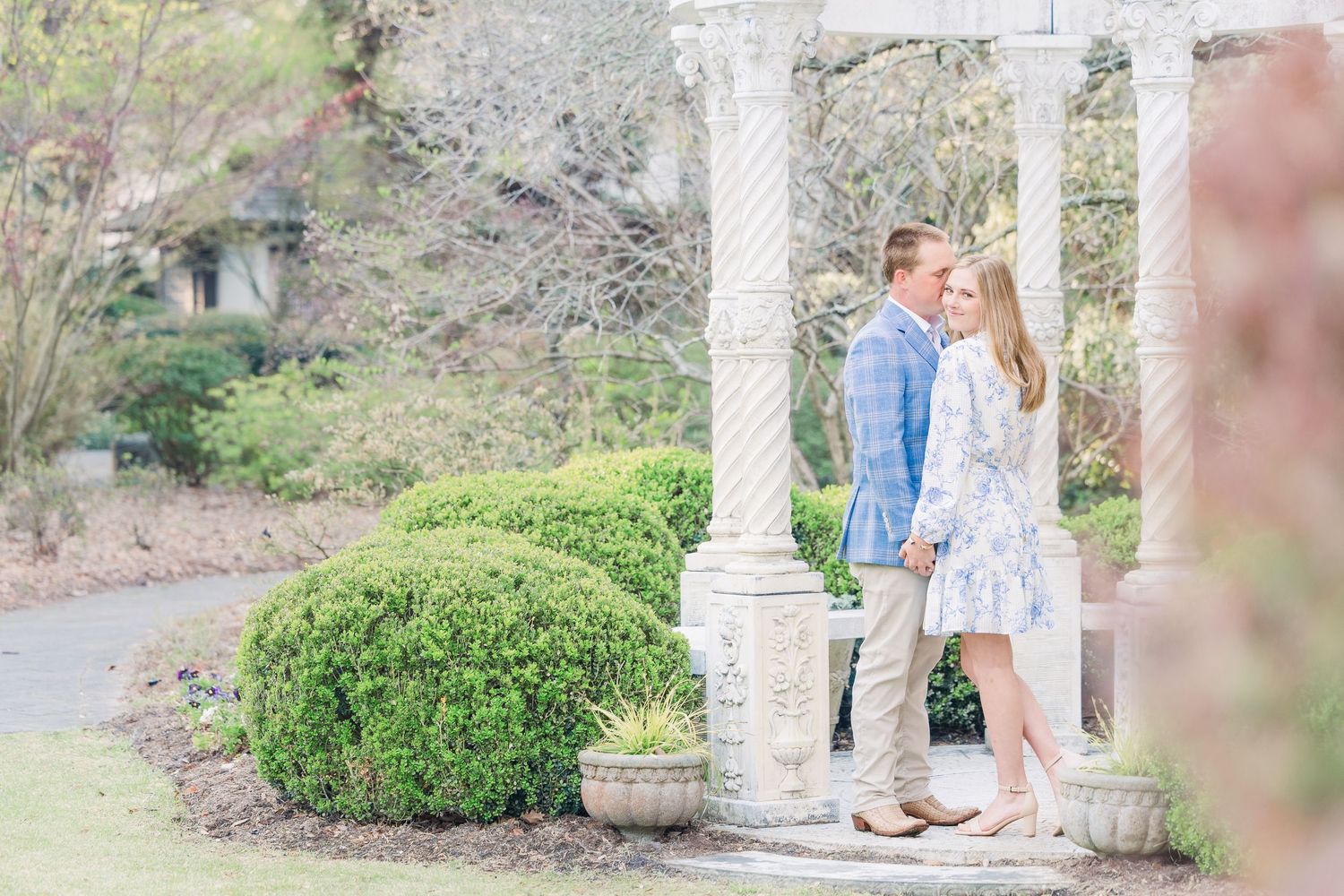 A romantic couple shares a tender moment in a garden setting with white columns and lush greenery.