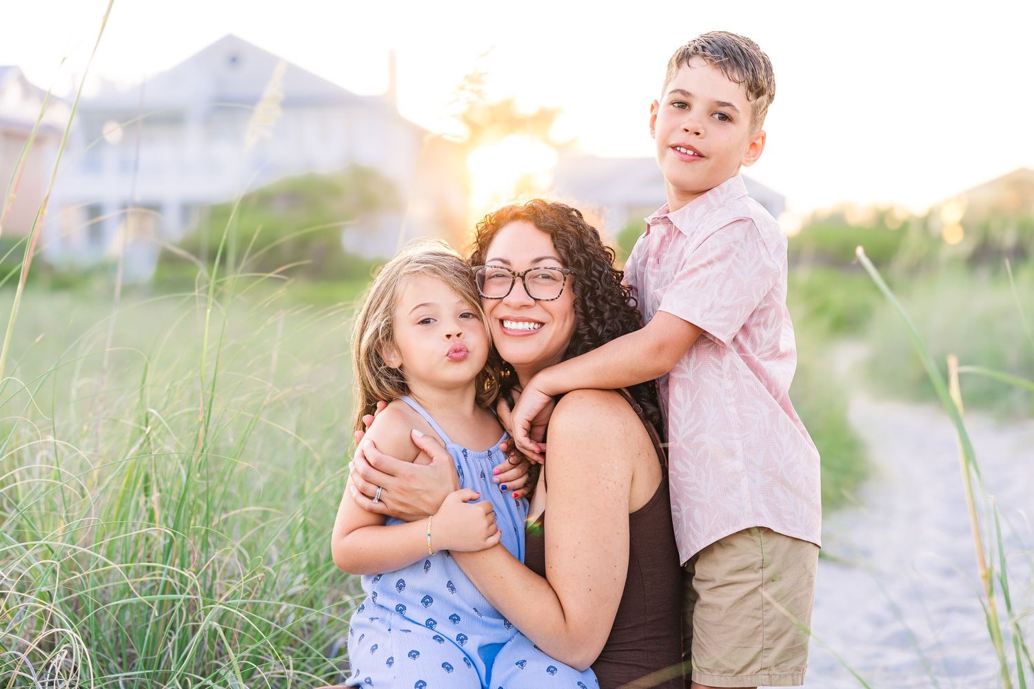 A mother smiles warmly while hugging her two young children on a sunny beach path surrounded by tall grass.