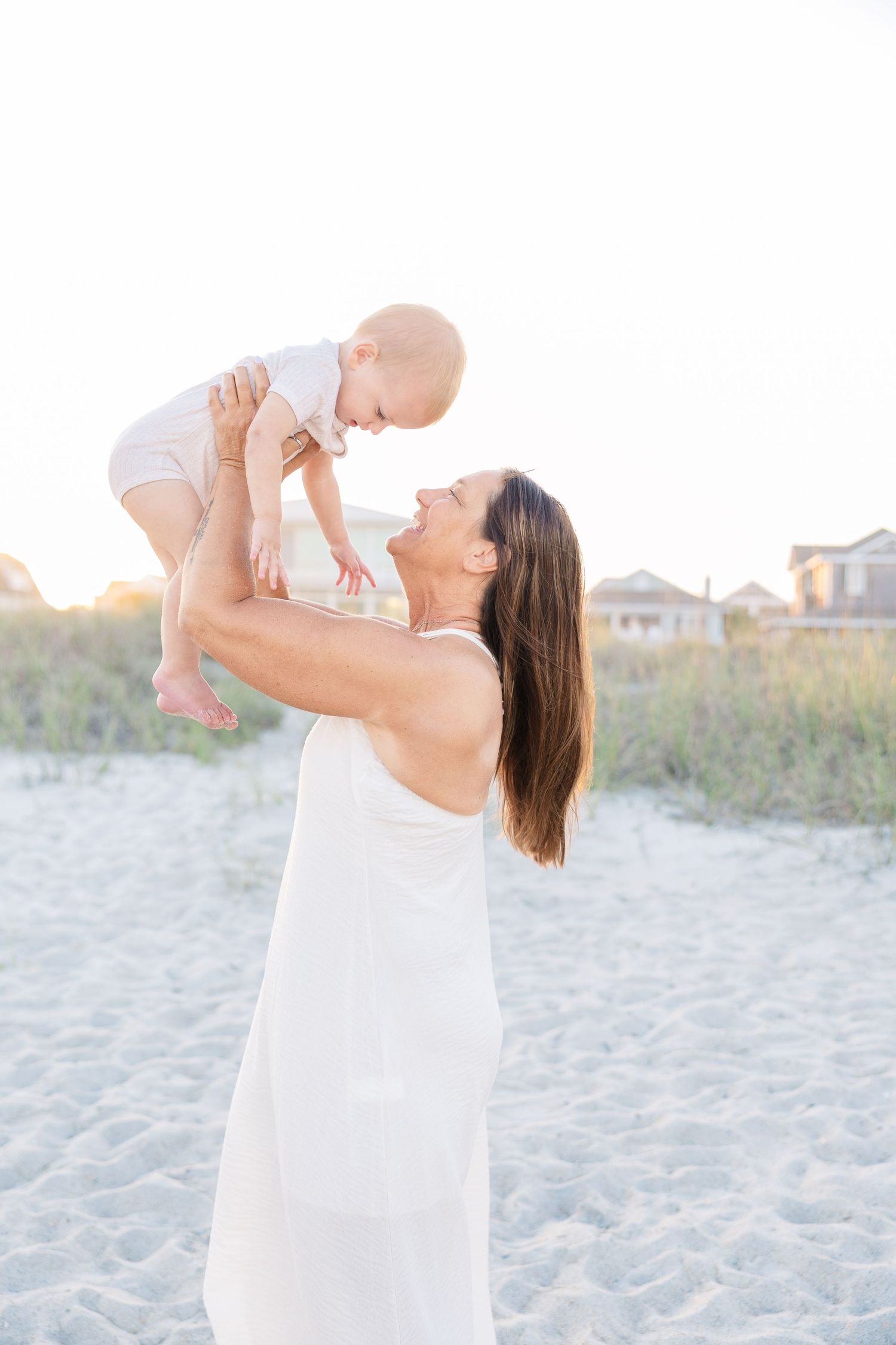 Joyful woman in white dress lifts laughing baby overhead on a sunny sandy beach.