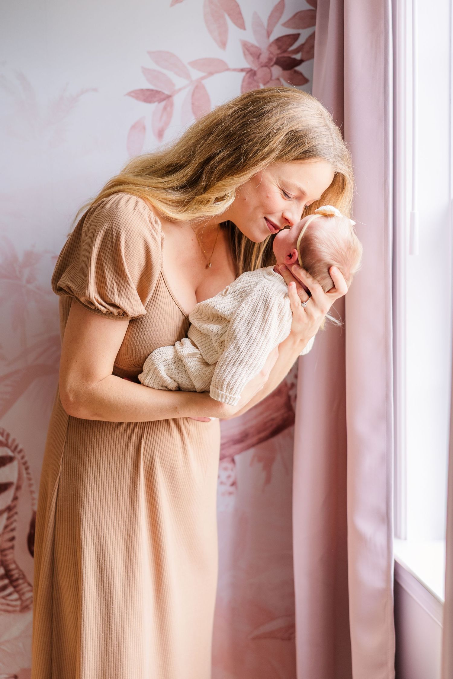 Tender moment between parent and newborn by window with pink floral wallpaper.