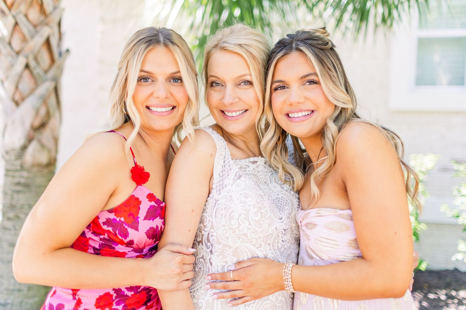 Three women in formal attire smile together for a close-up photo outdoors.