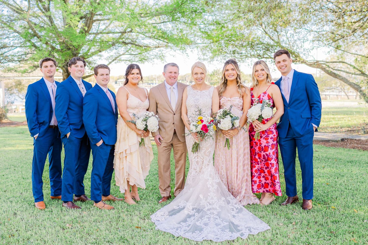 Full wedding party portrait with bridesmaids and groomsmen on sunny lawn with trees in background.