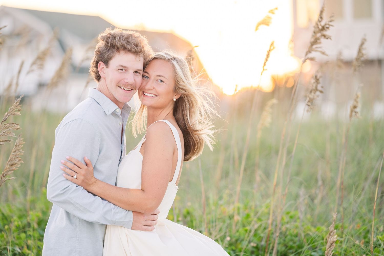 Romantic couple embracing on beach at sunset with soft lighting and coastal vegetation in background.